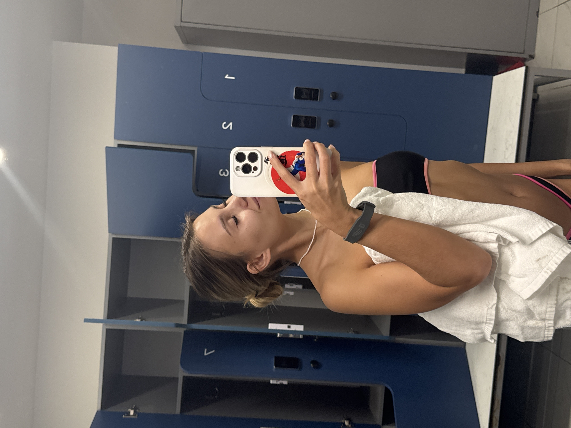 A woman in a black swimsuit with pink trim stands in a locker room. She is holding a cell phone up to take a selfie. She has a white towel draped over her shoulder. The locker room has blue lockers with silver handles. The numbers "1", "3", and "5" are visible on the lockers. The scene appears to be indoors and well-lit. The location, Chaikovsky, Russia, is not discernible from the image.