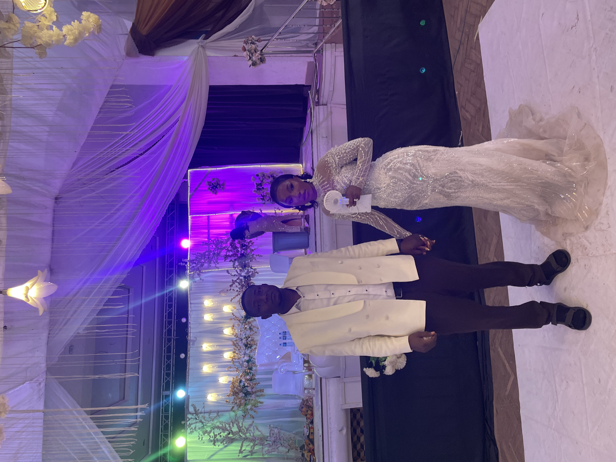 The image depicts a wedding scene in what appears to be an indoor venue in Jos, Nigeria. A bride in a long-sleeved, sequined white gown holds a small white fan. A groom in a cream-colored suit jacket and dark pants stands beside her, holding her hand. The background includes draped white fabric, lighting in various colors, and floral decorations. A dark backdrop is also present. The floor appears to be white or light-colored.