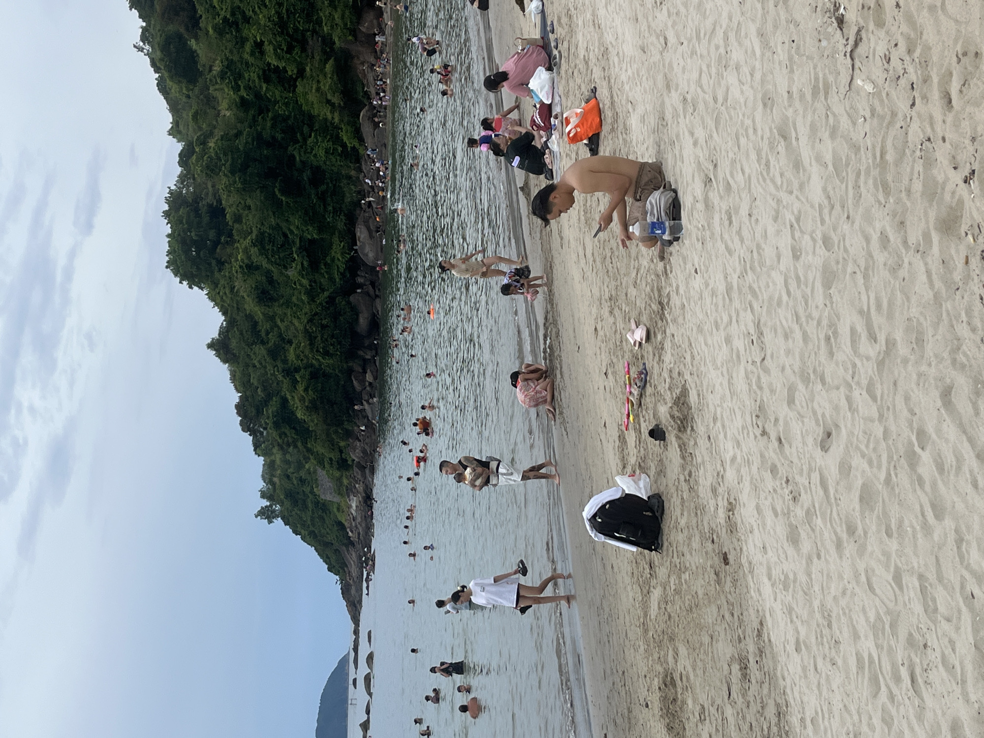 The image depicts a crowded beach scene at what appears to be a tourist destination in Da Nang, Vietnam, likely during the day given the overcast sky. The foreground is dominated by sand, with scattered personal items like a backpack, flip-flops, and a water bottle. In the middle ground, people are wading and swimming in the ocean. A man is holding a baby, a woman in a white t-shirt is walking along the water's edge, and other individuals are engaged in various beach activities. The background features a lush, green, tree-covered hillside that slopes down to the rocky coastline, characteristic of many beaches in the Da Nang region. The sea is calm with gentle waves. The overall atmosphere suggests a popular recreational spot with many visitors enjoying the water and the beach.