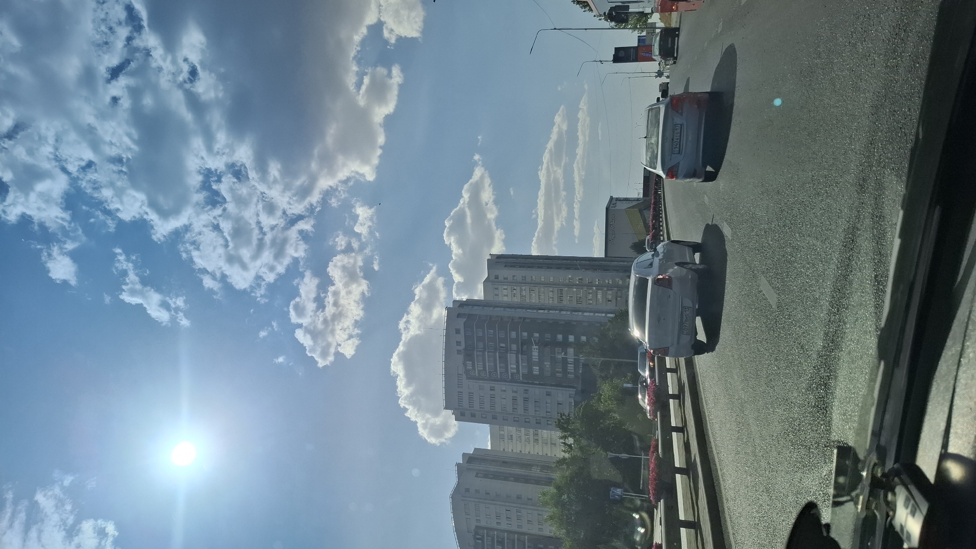 The image captures a sunny day in Shymkent, Kazakhstan, likely on a highway or a major city road. The sky is a vibrant blue, dotted with fluffy white clouds, and the sun shines brightly, creating lens flare on the left side of the frame. In the background, several tall, modern apartment buildings are visible, characterized by their uniform windows and geometric architecture.
The foreground is dominated by a multi-lane asphalt road where several cars are in motion. The perspective suggests the photo was taken from inside a vehicle, looking forward. A white sedan is in the left lane, and a silver sedan is in the right lane, both moving away from the camera. Further down the road, other vehicles are also visible, indicating moderate traffic. The sides of the road are lined with green trees and a median with red flowers. The overall scene depicts a typical urban environment during daylight hours in Shymkent.