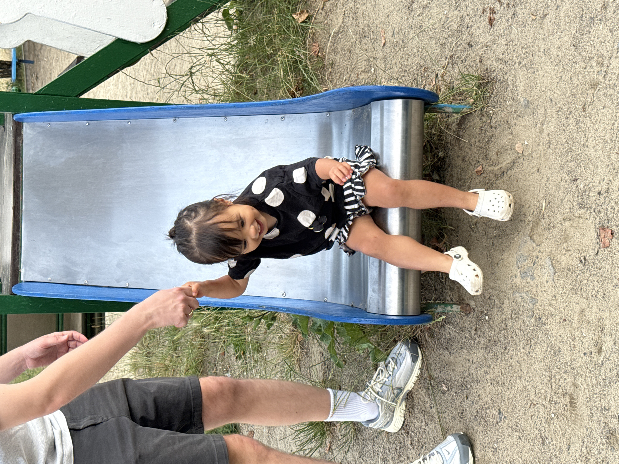 A young child with dark hair, wearing a black and white polka dot shirt and a ruffled black and white striped skirt, sits on a metal slide at a playground. The child is smiling and holding onto the hand of an adult, who is wearing grey shorts and a light-colored t-shirt. The adult's hand is visible, and they are wearing a gold ring. The playground appears to be located outdoors with a sandy ground and grass. The slide has blue metal edges. The scene is likely in Lutsk, Ukraine, based on the provided information.