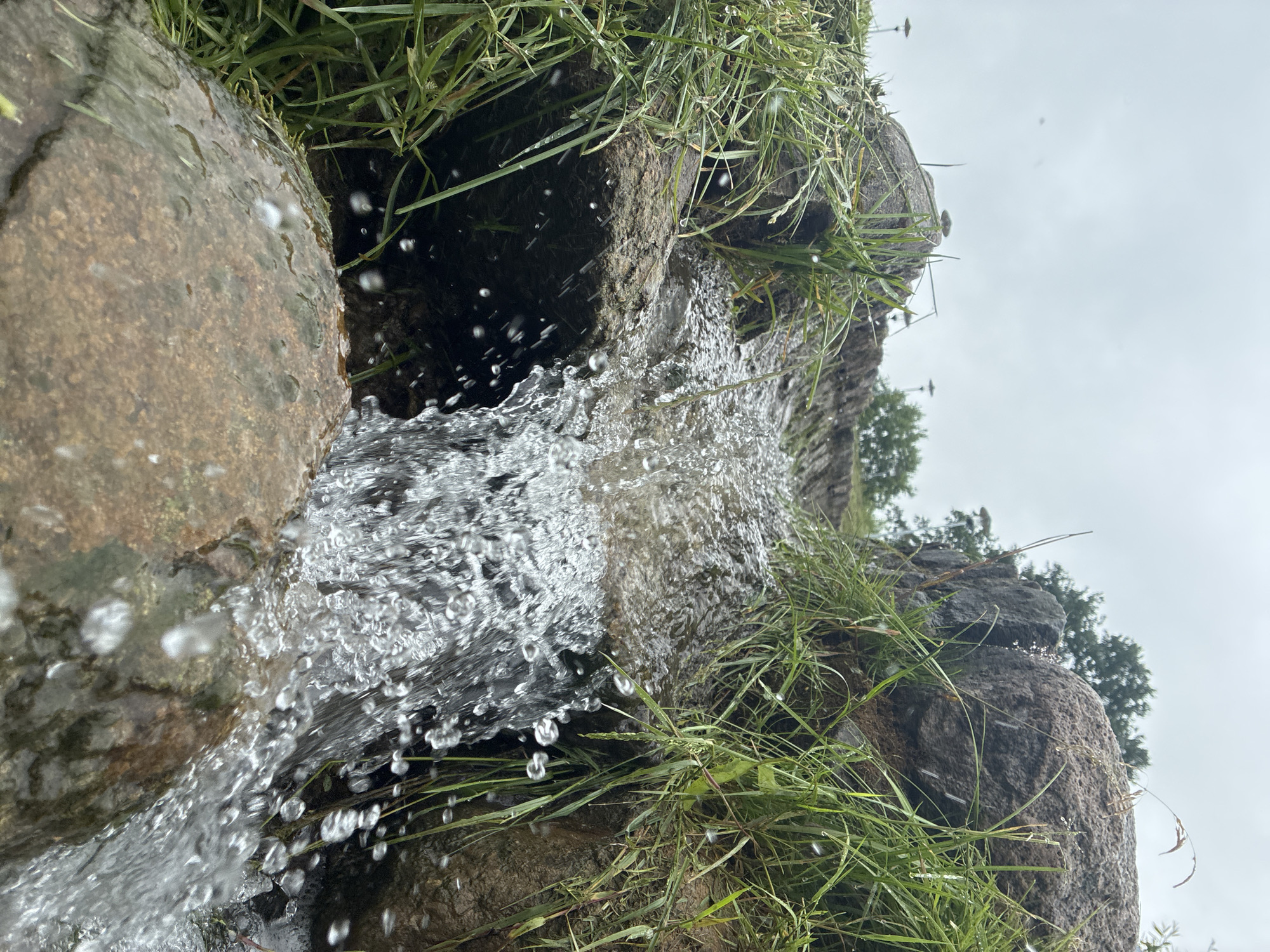 The image shows a close-up of a small waterfall flowing over rocks and grass. The water is clear and appears to be in motion, with droplets visible in the air. Green grass and brown rocks surround the waterfall. The sky is overcast and appears to be in the background. The location is identified as Sabile, Latvia. There are no people or animals visible in the image.
