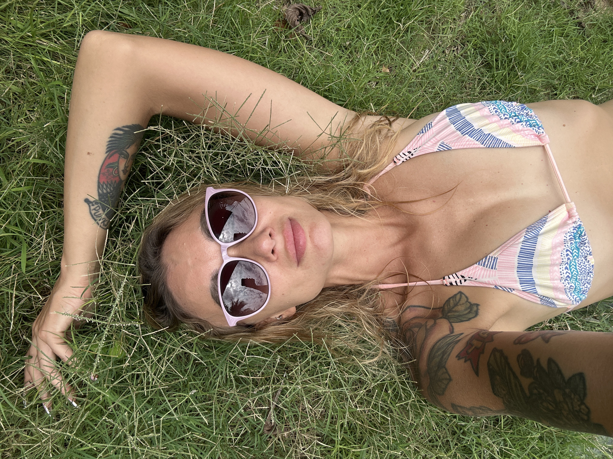 A young woman is lying on her back in green grass, viewed from a high angle looking down. She has blonde or light brown hair spread around her head and is wearing a colorful bikini top with patterns in pink, blue, yellow, and white. Her eyes are covered by light purple-framed sunglasses with mirrored lenses, which reflect a bright, cloudy sky and what appears to be a palm tree. Her lips are slightly parted, revealing light pink lipstick.
She has several tattoos on both arms. On her left forearm, a distinct tattoo of a stylized head with a mohawk is visible, with the word "PUNK" written below it. Other tattoos, appearing to be floral or nature-themed, are visible on her upper left arm and her right arm. Her left hand is resting in the grass, fingers slightly curled. The grass itself is green but somewhat sparse in places, with a few dry leaves scattered around. The overall scene suggests a bright, sunny day, indicating outdoor relaxation. The location is Mũi Né, Vietnam.