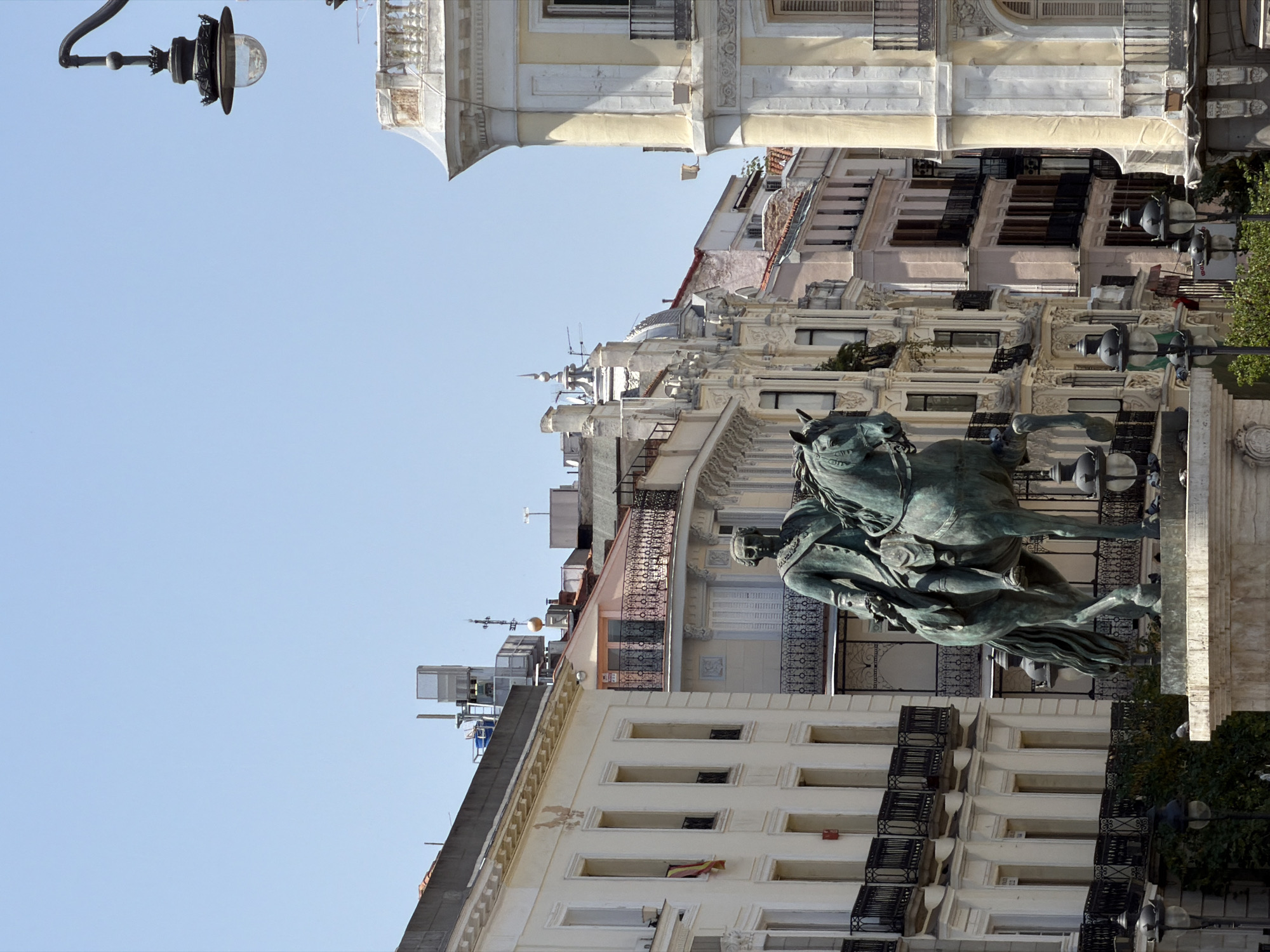 This is a medium shot, eye-level, outdoor, daytime photograph of a statue of a man on horseback in a city square. The statue is made of a dark green metal and depicts a man in a military-style jacket and breeches. He is seated on a horse that is depicted in mid-stride, with its front legs raised. The statue is on a large stone pedestal. The statue is located in Puerta del Sol in Madrid, Spain, as evidenced by the surrounding architecture.
The buildings in the background are a mix of architectural styles, with ornate facades, balconies with black wrought-iron railings, and arched windows. The buildings are painted in light colors, such as cream and pale yellow, with some featuring terracotta-tiled roofs. The sky is clear and bright blue. A street lamp with a dark metal frame and a glass globe is visible in the upper right corner of the image. There are several other street lamps visible at the base of the statue, along with some pigeons on the pedestal. The overall scene is one of a historic urban environment on a sunny day.
