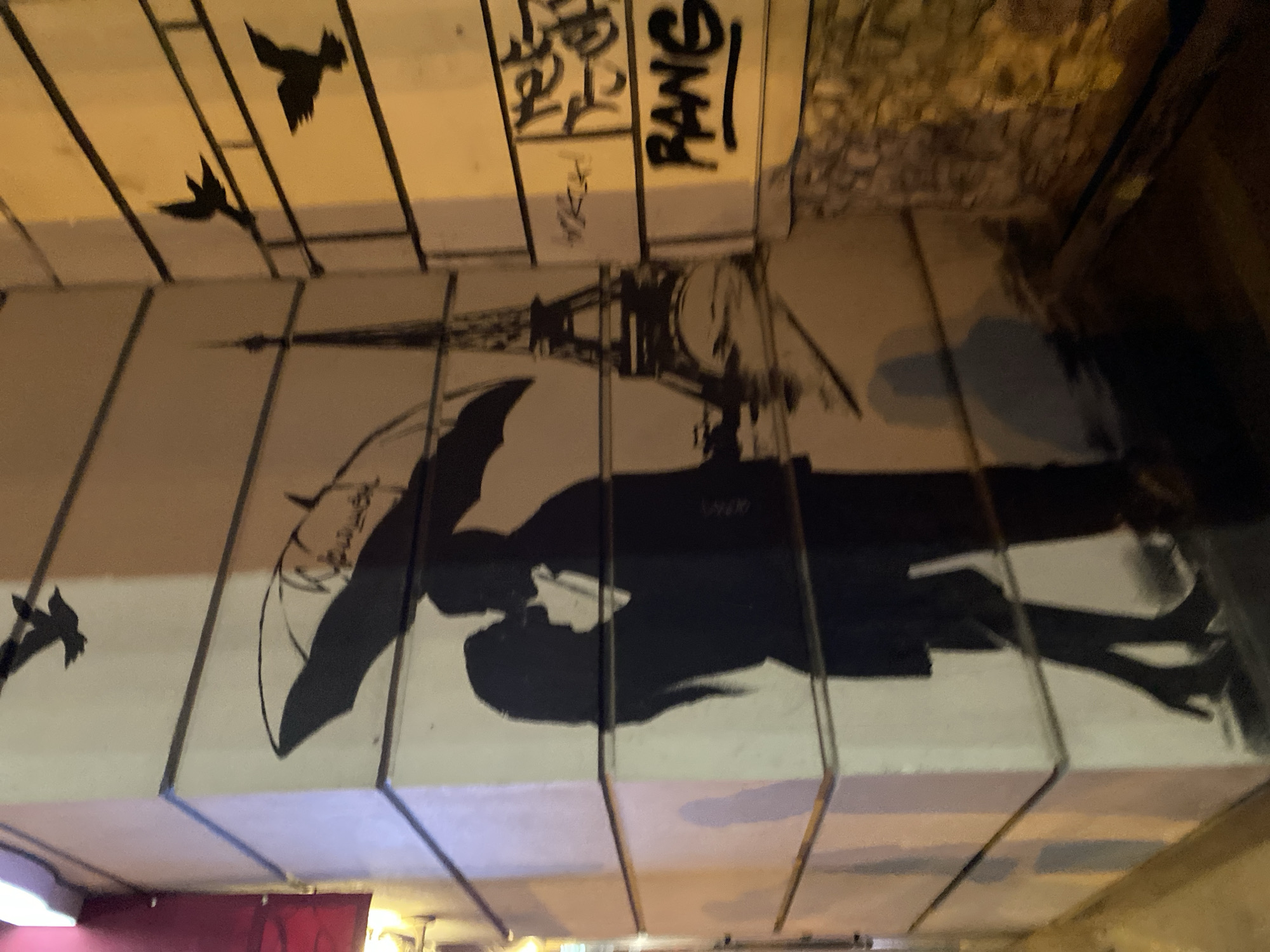The image shows street art located in Yekaterinburg, Russia. The mural depicts a couple kissing under an umbrella, with the Eiffel Tower in the background. There are bird silhouettes painted on the wall near the top left corner of the image. Graffiti is present in the upper right corner. The painting is applied to rectangular concrete blocks that form part of a structure, likely a bridge or underpass. The lighting is dim, possibly at night.