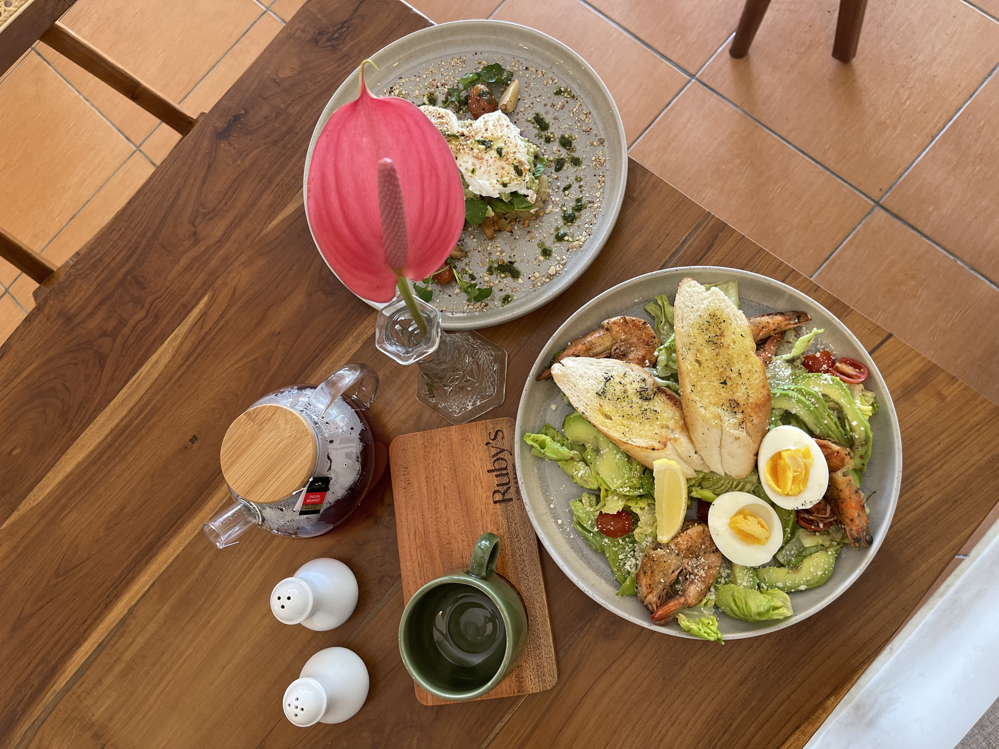 A top-down shot on a wooden table showcases two plates of food, a teapot, salt and pepper shakers, and a small green mug on a wooden coaster. The food on the left plate appears to be a salad with shrimp, hard-boiled eggs, avocado, and toasted bread. The plate on the right contains what looks like poached eggs on a bed of vegetables, garnished with a vibrant pink flower. The setting suggests a meal in Kuta, Indonesia, likely at a cafe or restaurant with outdoor seating, given the tiled floor and wooden furniture. The overall atmosphere is casual and relaxed.