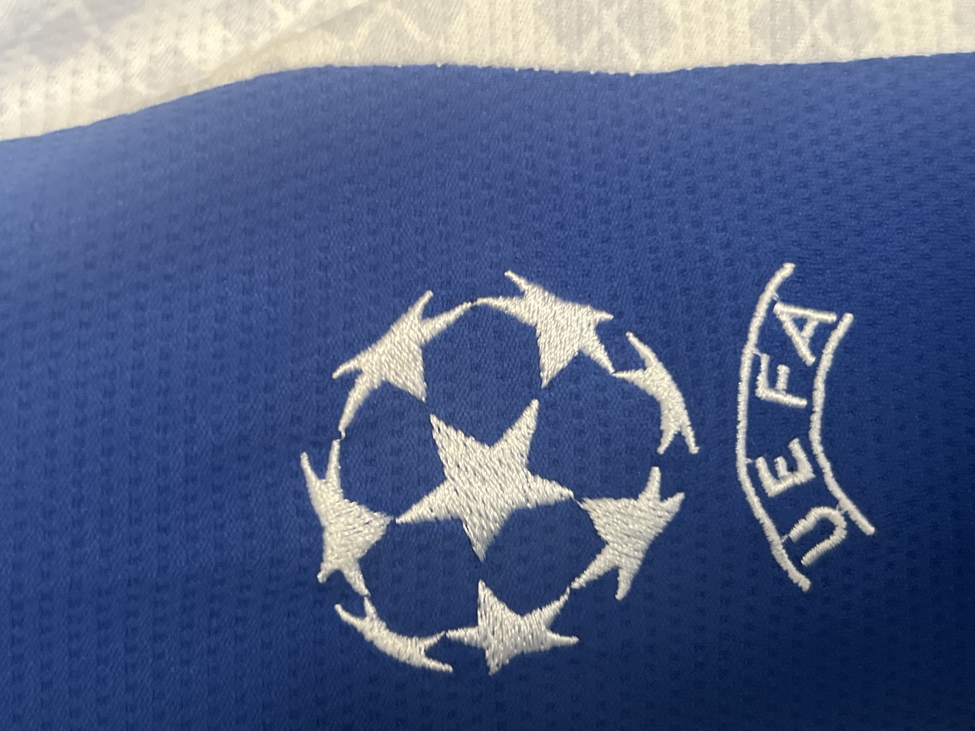 The image shows a close-up of a blue fabric with white embroidered designs. The main design is the UEFA Champions League logo, a stylized football made of white stars. Below the logo, the letters "UEFA" are also embroidered in white, forming an arc. The fabric appears to be a mesh material, typical of sportswear. The background to the right of the blue fabric is a blurred white material, possibly another piece of clothing or a surface. The image does not provide any information about the location in the city of Jauro Yinu, Nigeria, or any people, actions, or interactions.