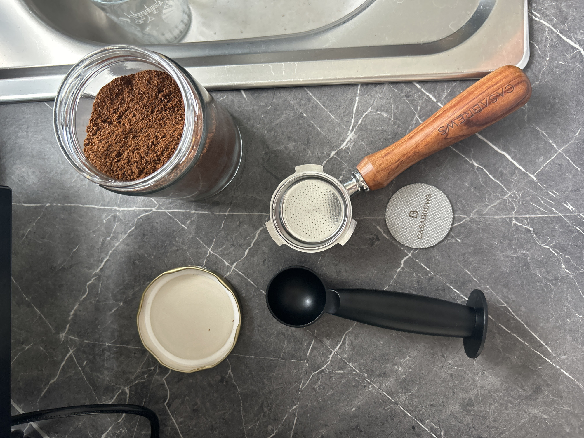 The image shows a collection of coffee-related items arranged on a gray countertop with white marble-like veining. A glass jar containing ground coffee sits to the left of a portafilter with a wooden handle branded "CASABREWS". A gray circular filter with "CASABREWS" printed on it is next to the portafilter. Below are a black coffee scoop and the lid of the jar. The top of a stainless steel sink or tray is visible in the background. The environment suggests a domestic kitchen setting. There is no indication of a location in London, United Kingdom.