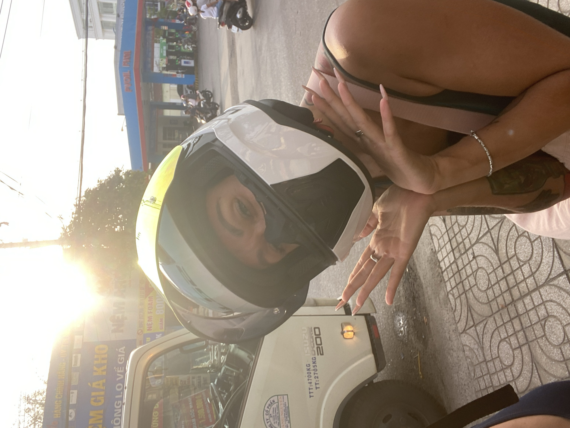 A woman wearing a white, yellow, and black motorcycle helmet with the visor down is posing for a photo. She is holding her hands near the helmet, showing off long fingernails and a silver ring. Her left arm is visible, with a silver bracelet and a tattoo. In the background, a white Isuzu truck with Vietnamese writing and "200" is partially visible. Beyond the truck, there are signs and a gas station with the name "PVOIL FICT" visible. Further in the background, there are motorcycles and people. The location is Phan Thiết, Vietnam, based on the Vietnamese writing. The lighting suggests it is late afternoon or early morning due to the sun's position. The ground is paved with patterned tiles.