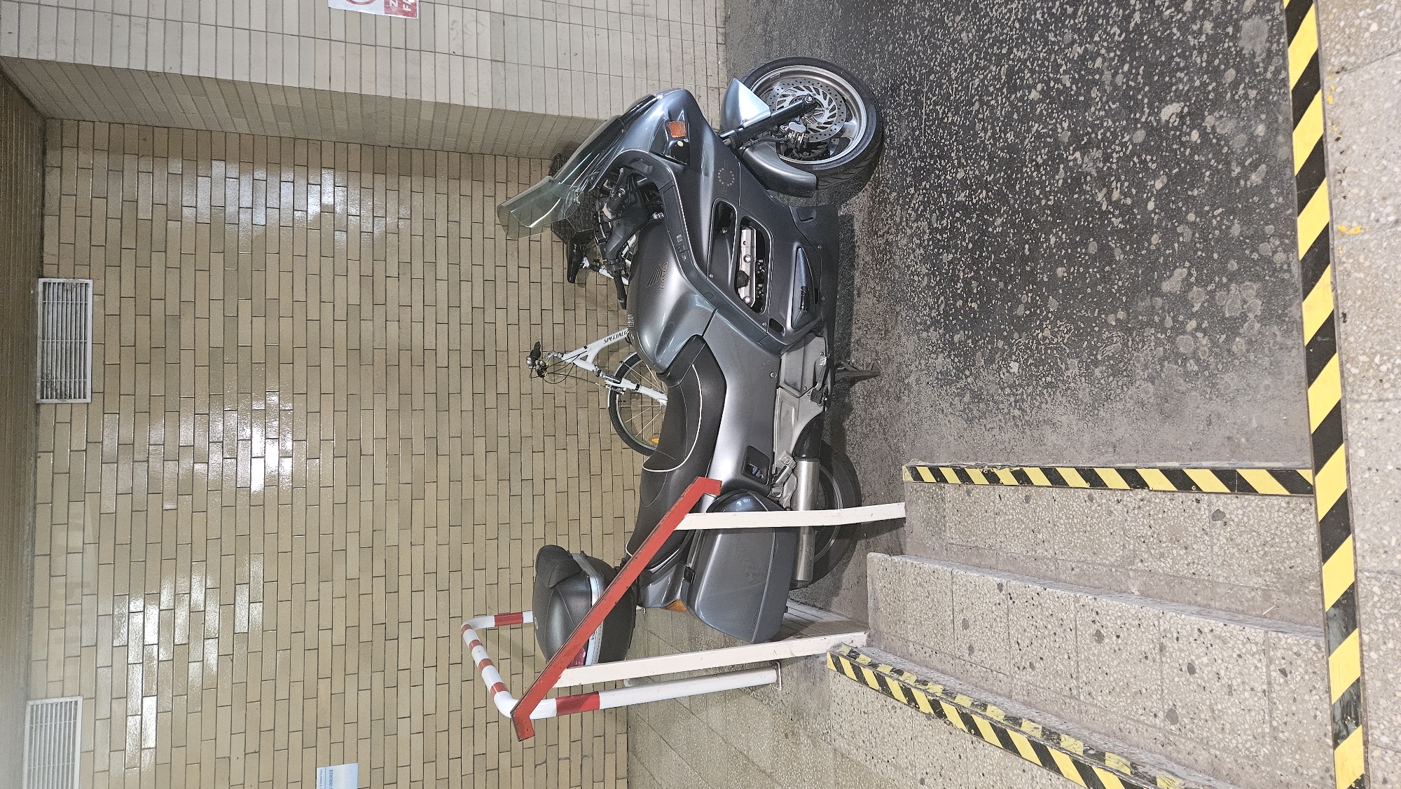 The image depicts an outdoor area with a beige brick wall in the background. Two white bicycles are parked behind a gray motorcycle. The motorcycle is a touring model with saddlebags and a windshield. A red and white barrier is positioned in front of the motorcycle. The ground surface is a mix of concrete and asphalt, marked with yellow and black hazard stripes on steps and at the edge of the ground. The scene appears to be in a utilitarian or public space, possibly a parking area or a section of a building. There are no people visible in the image.