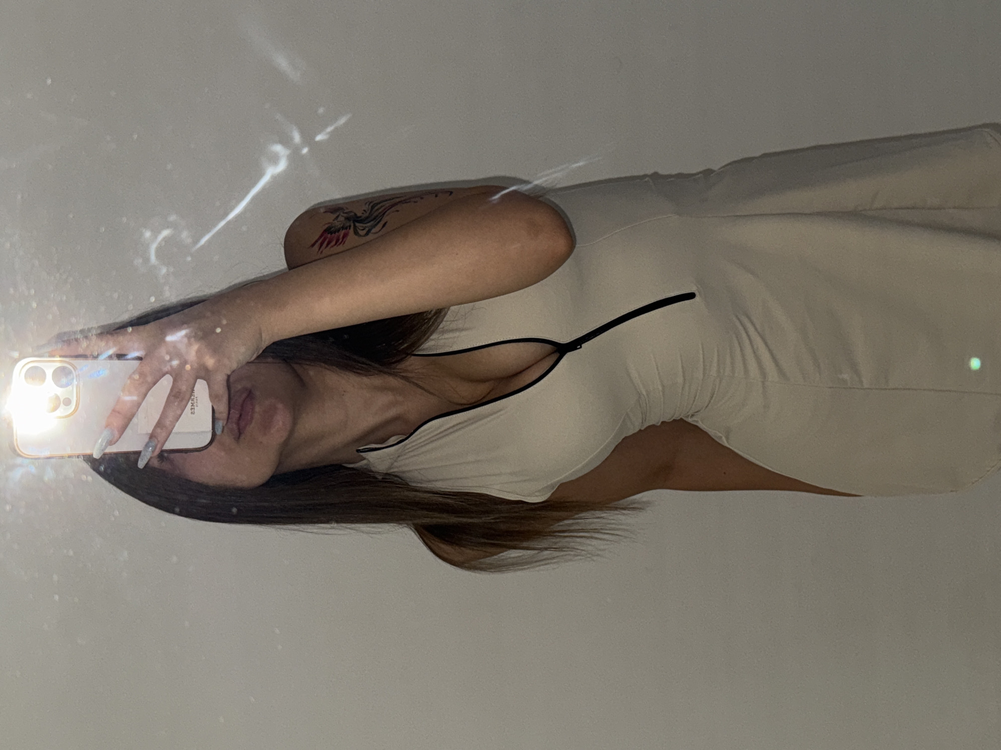 A woman with long, dark hair and light-colored nails is taking a mirror selfie in a room with a plain, light-colored wall. She is wearing a sleeveless, light-colored dress with a black zipper down the front. A tattoo of a bird is visible on her upper left arm. The phone she is holding is partially obscuring her face. The lighting in the room creates some reflective glare on the wall. There is no indication that this scene is located in the city of Sofia, Bulgaria.