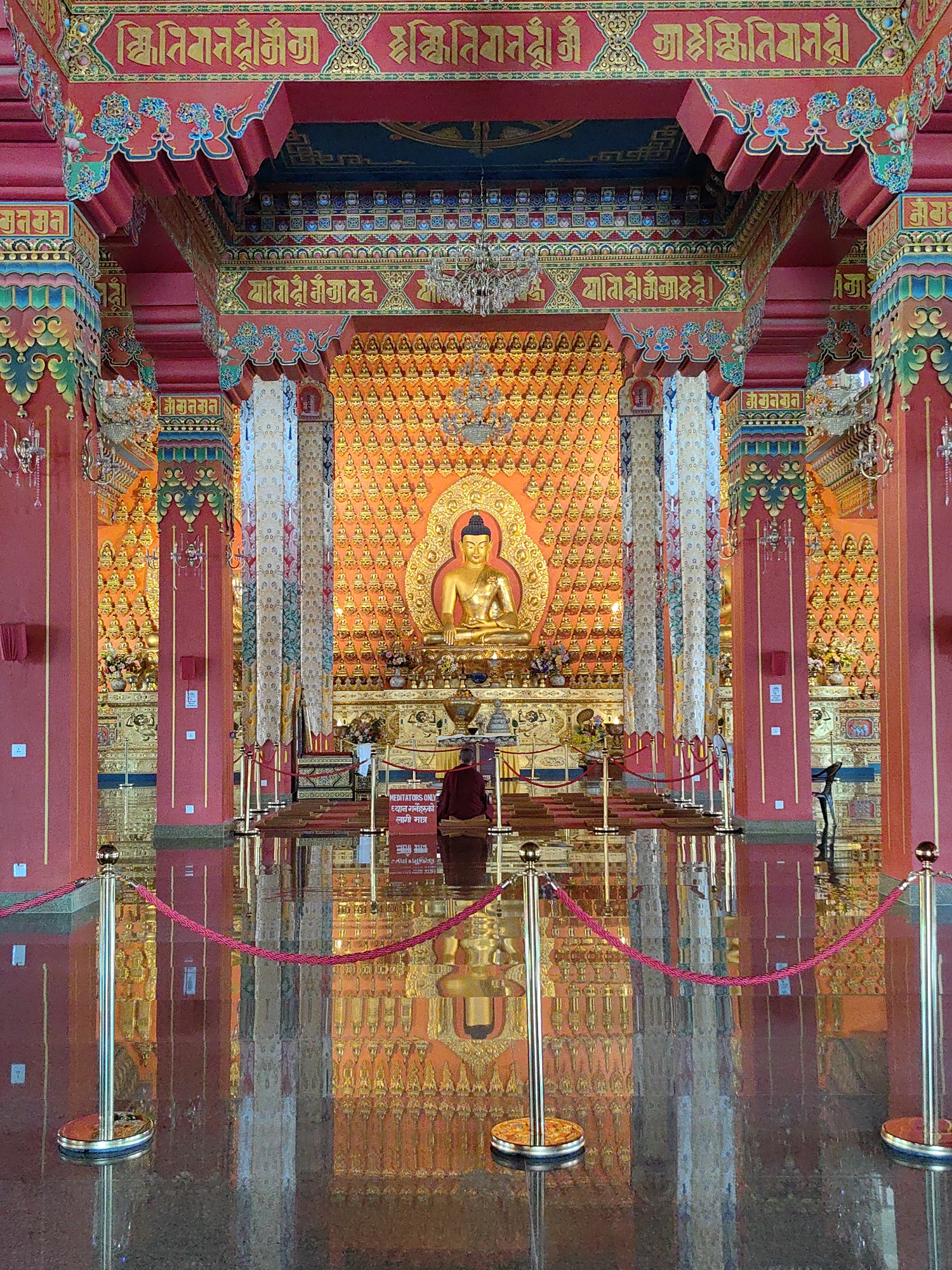 The image depicts the interior of a Buddhist temple, likely the Great Buddha Temple in the city of Parsa, Nepal, given its ornate architectural style. A large, golden statue of the Buddha is centrally positioned against a backdrop filled with numerous smaller golden Buddha effigies. The temple is richly decorated with red and gold, featuring intricate patterns and Tibetan script on the pillars and ceiling. Red velvet ropes on golden stanchions guide visitors, and a reflective, polished floor mirrors the opulent surroundings. A lone figure, dressed in maroon robes, is seated in meditation facing the main Buddha statue, suggesting a place of worship and contemplation. The atmosphere appears serene and sacred, illuminated by the warm glow of the golden statues and decorative elements.
