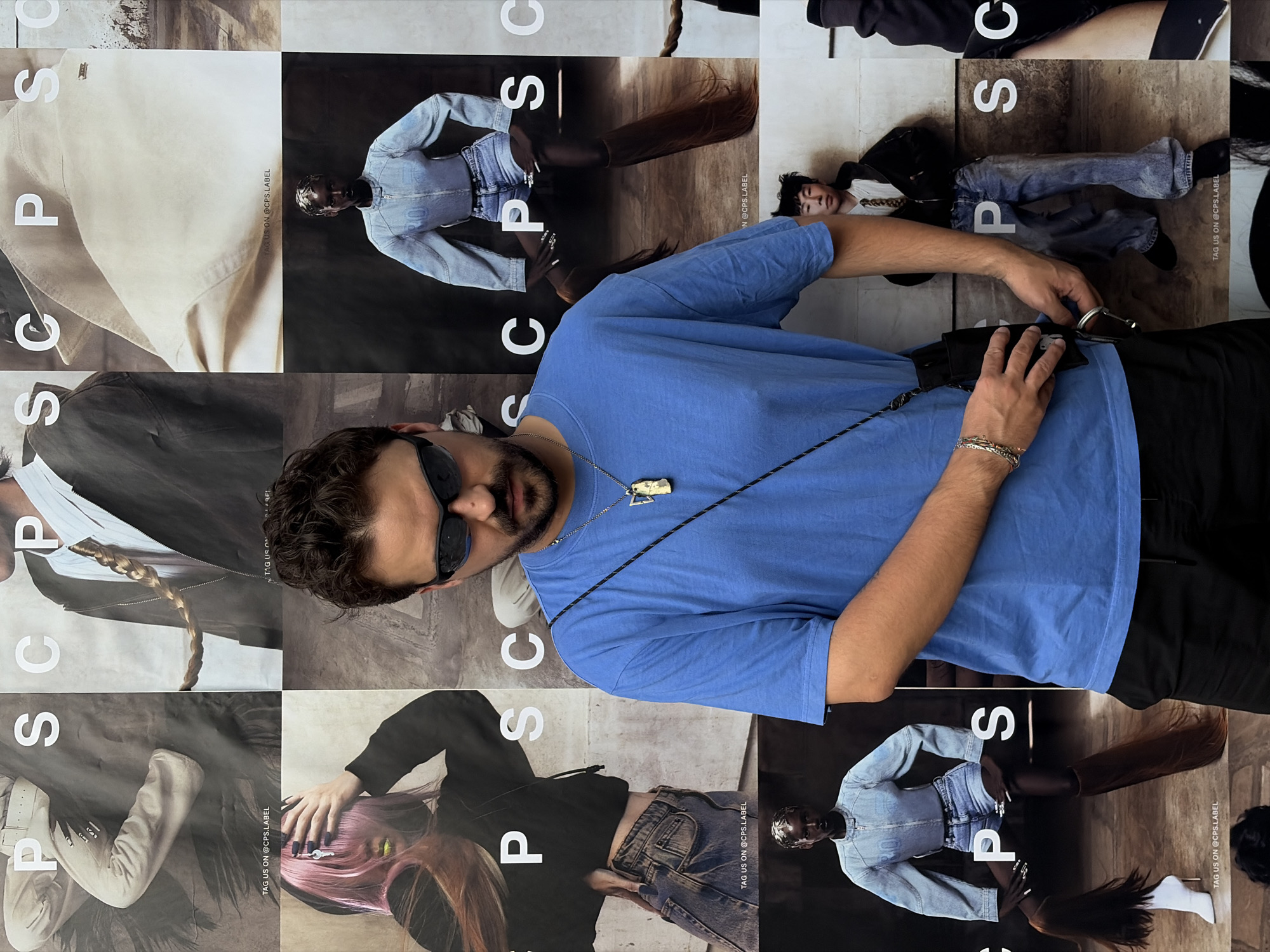 The image shows a man with dark curly hair and sunglasses wearing a blue T-shirt, black pants, a necklace, and a wrist bracelet. He is standing in front of a wall made up of multiple fashion magazine pages with the brand name "PSC" printed on them, along with the phrase "TAG US ON @CPS.LABEL". The backgrounds of the magazine pages vary, featuring different models wearing fashionable clothing. The scene is not explicitly set in Bangkok, Thailand.