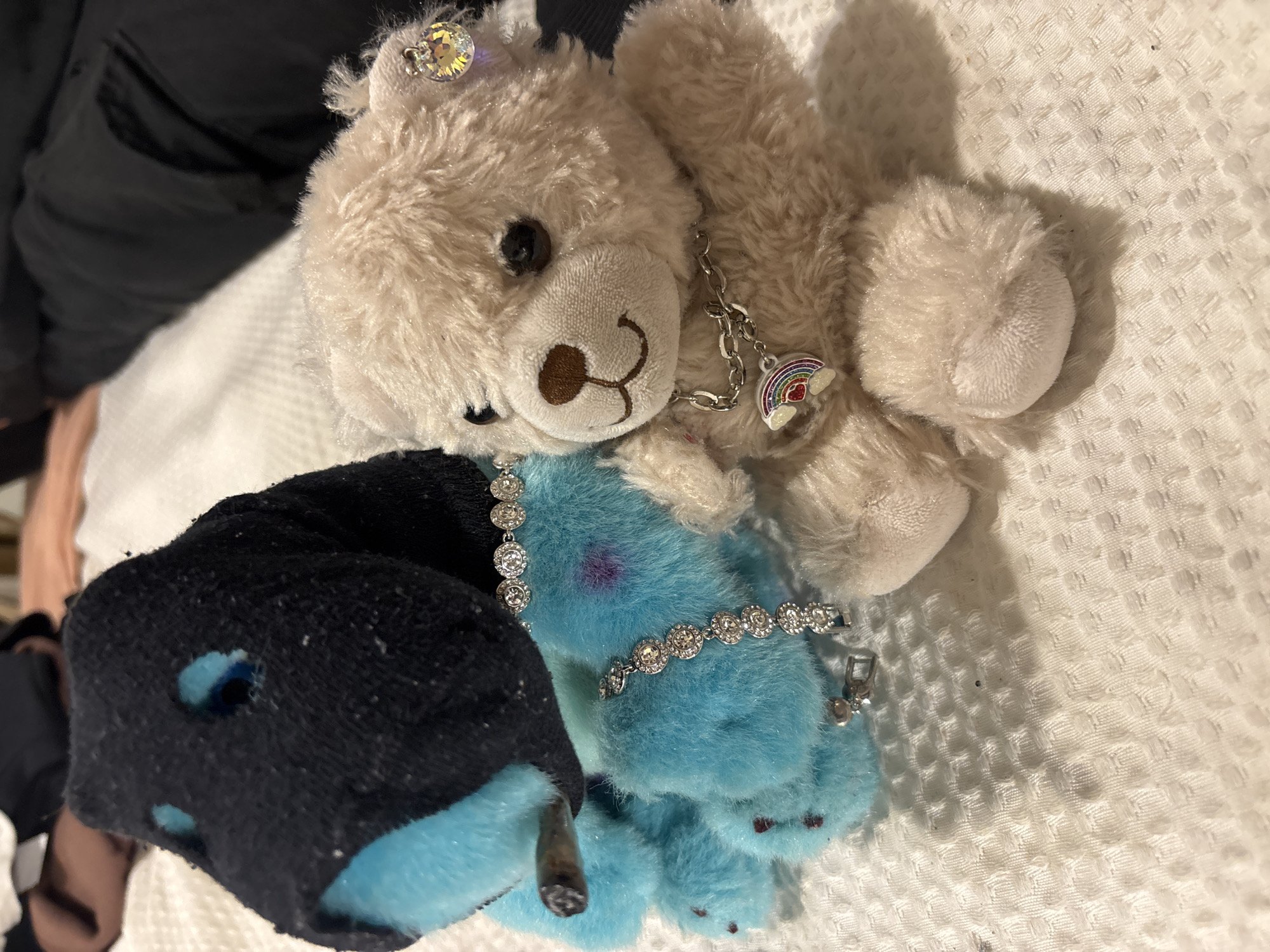 A close-up shot displays three plush toys arranged on a white, textured surface. A light brown teddy bear, adorned with a jewel on its head and a necklace with a rainbow charm, lies atop a blue bear which is wearing a jeweled bracelet. A black sock with holes is positioned on the left side of the frame. The location is not discernible from the image; therefore, there is no information available about its connection to Buenos Aires, Argentina. There is no evidence of people, actions, or interactions in the image.