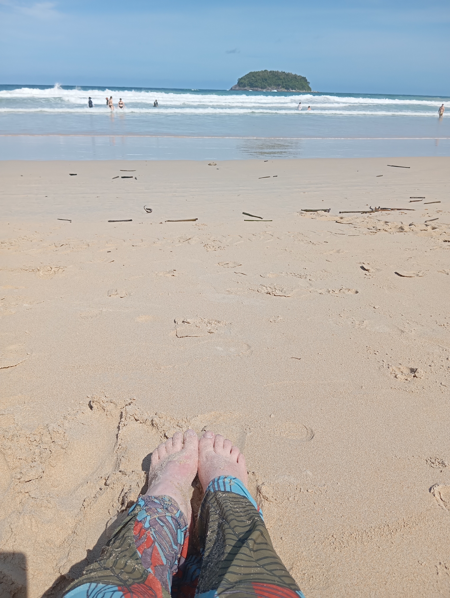 This is a photograph taken at Karon Beach in Karon, Thailand, looking out towards the ocean. The foreground is dominated by fine, light-colored sand, with the lower legs and feet of a person wearing colorful patterned pants submerged in it. Footprints are visible in the sand.
In the midground, the gentle waves of the ocean meet the shore, with white surf. Several people are visible in the shallow water, appearing to be playing or wading. Further out, on the horizon, a small, tree-covered island is visible, known locally as Koh Pu. The sky above is clear and blue with a few wispy clouds. The scene depicts a sunny day at the beach, with a sense of leisure and relaxation.