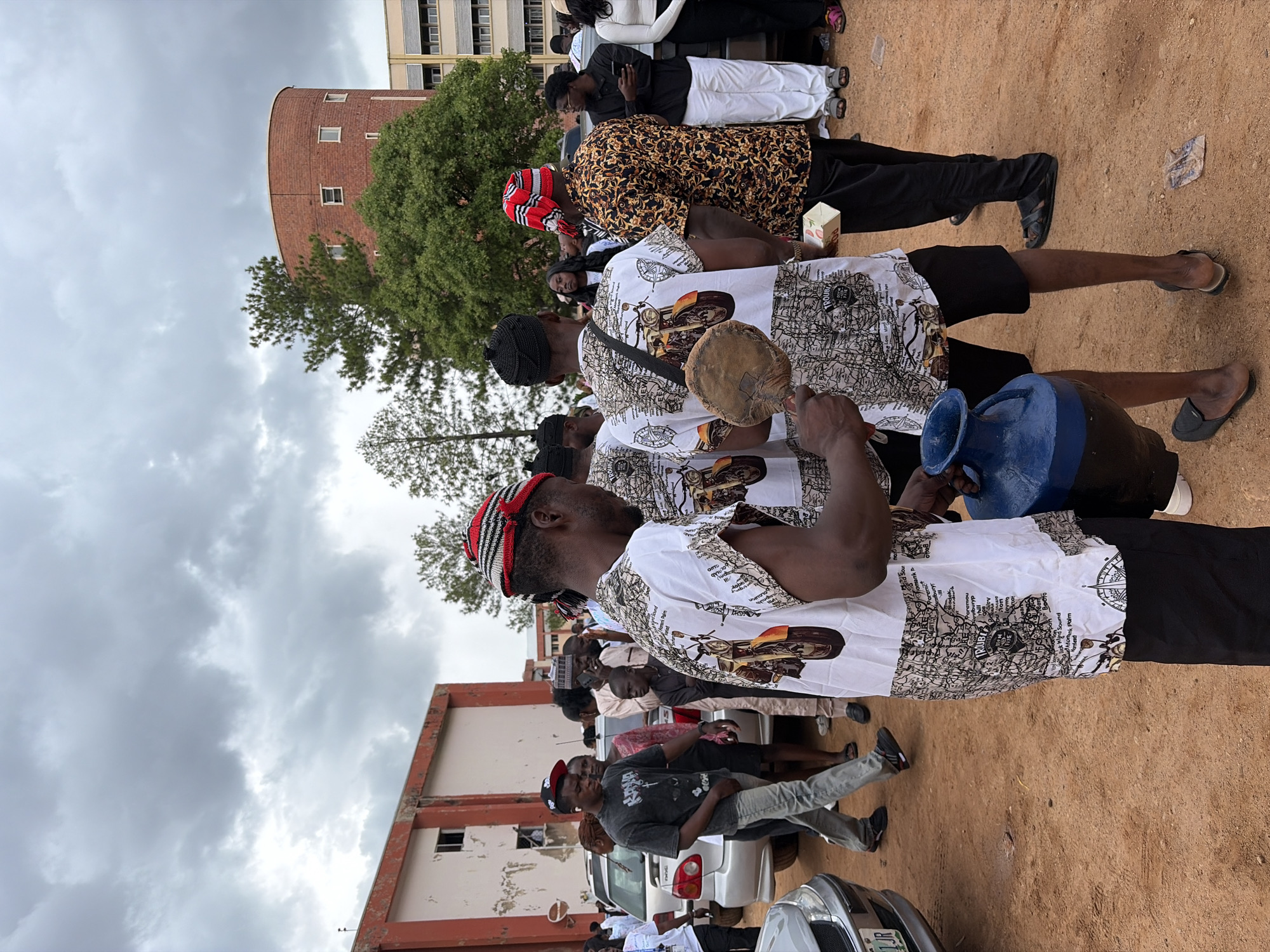 In the city of Jos, Nigeria, a group of young men in distinctive patterned shirts are gathered outdoors, with a backdrop of buildings and a cloudy sky. The shirts feature intricate designs, including what appears to be a map and motorcycles. Two individuals in the foreground are prominently featured, one holding a blue vase-like object and another holding a round, flat object resembling a drum beater. Other people are visible in the background, some standing near cars, and one man is wearing a red and black striped headwrap. The ground is a reddish-brown dirt. The overall atmosphere suggests a communal gathering or event.