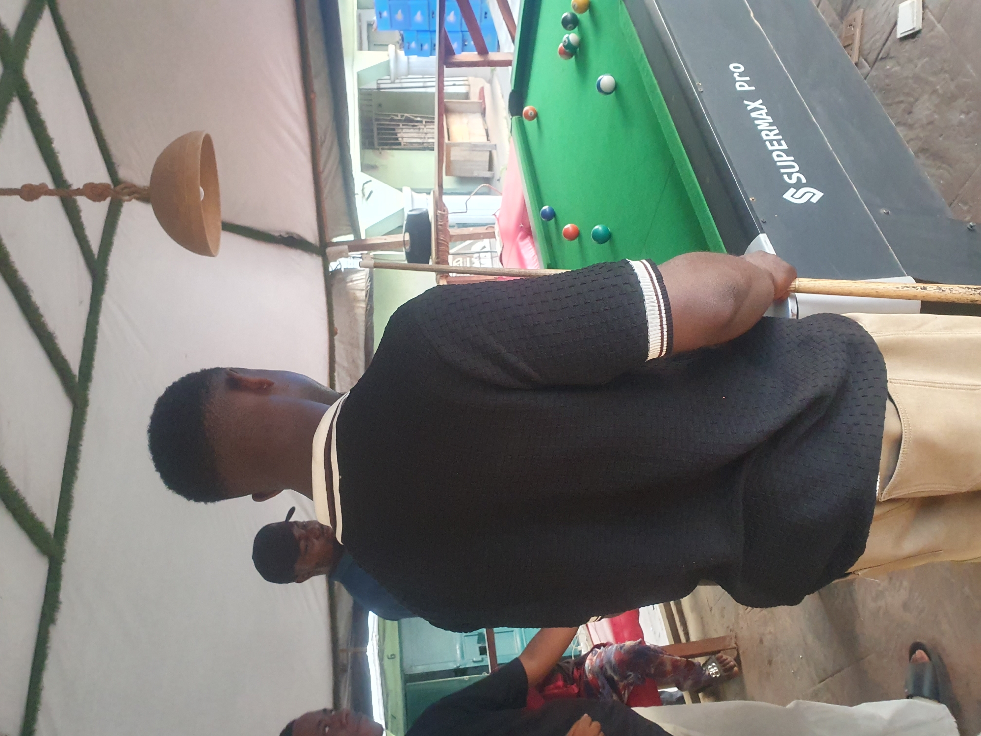 A man is playing pool on a "Supermax Pro" table. Several other people are standing around watching. The setting appears to be outdoors or under a temporary shelter, possibly in Lokoja, Nigeria, although there is nothing to confirm this.