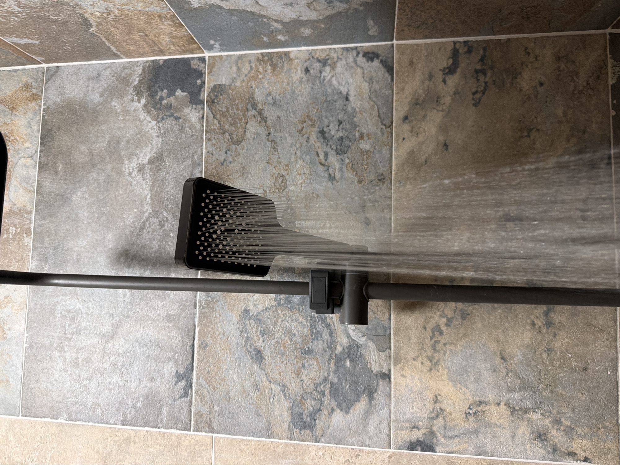 A close-up, eye-level shot shows a modern, dark-colored shower head with water actively spraying from it, creating a misty effect. The shower head is mounted on a dark metal pole, which extends downwards out of frame. The background is composed of textured, earth-toned tiles, suggesting a bathroom environment. The tiles have a natural stone appearance with variations in color and pattern, ranging from light beige to dark brown and gray. The lighting is consistent throughout the image, illuminating the shower and tiles without harsh shadows. There are no people visible in the frame. The image does not provide any information or visual cues that would allow for the determination of its location in Sofia, Bulgaria.