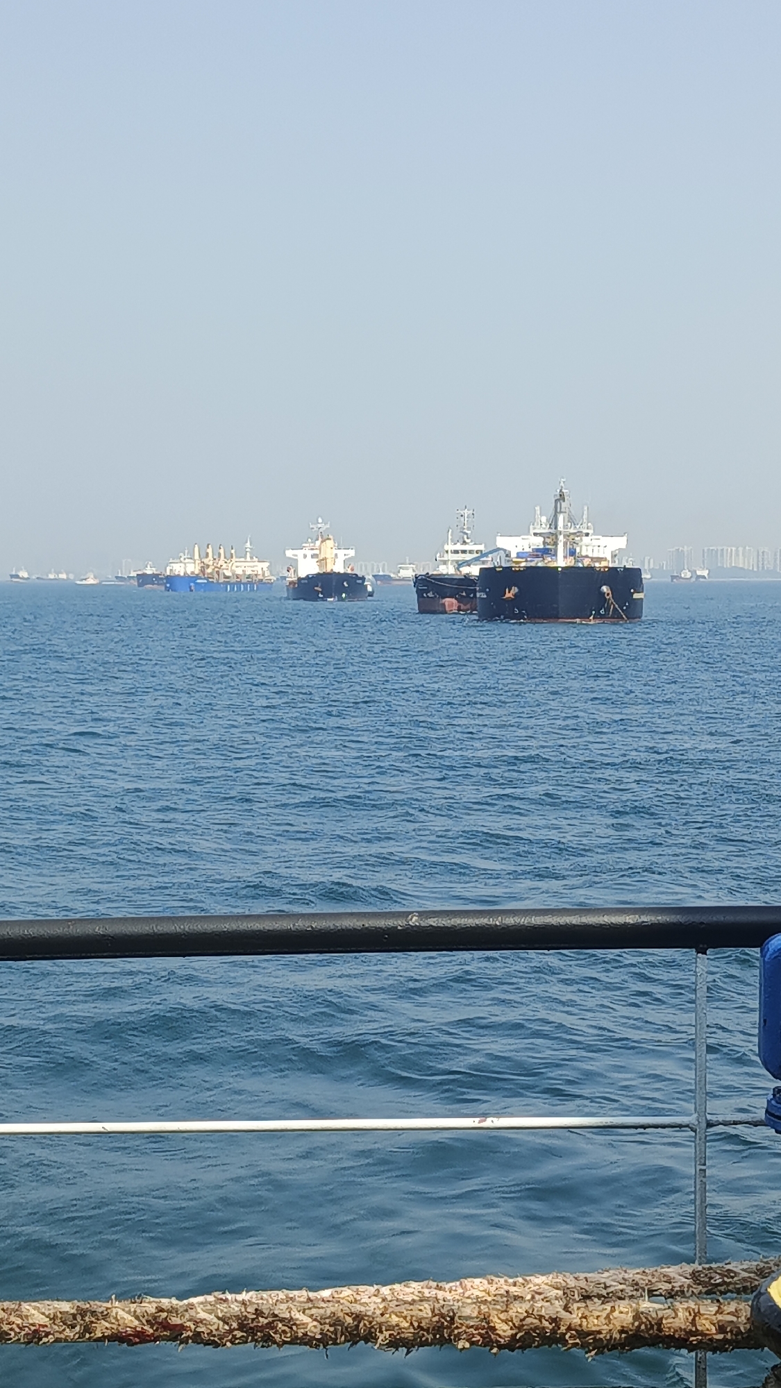 A wide-angle, portrait-oriented, eye-level outdoor shot shows a hazy, bright sky over a calm, blue ocean. Multiple large cargo ships of various sizes are anchored and appear to be awaiting port. In the foreground, a black railing runs horizontally across the bottom of the image, with a thick, woven rope visible below it. The ships are the primary focus, with several appearing as silhouettes against the distant, hazy, urban skyline which may indicate the location as Singapore. The water is a deep blue with small ripples, suggesting minimal wave action. The overall impression is one of a busy maritime port environment.