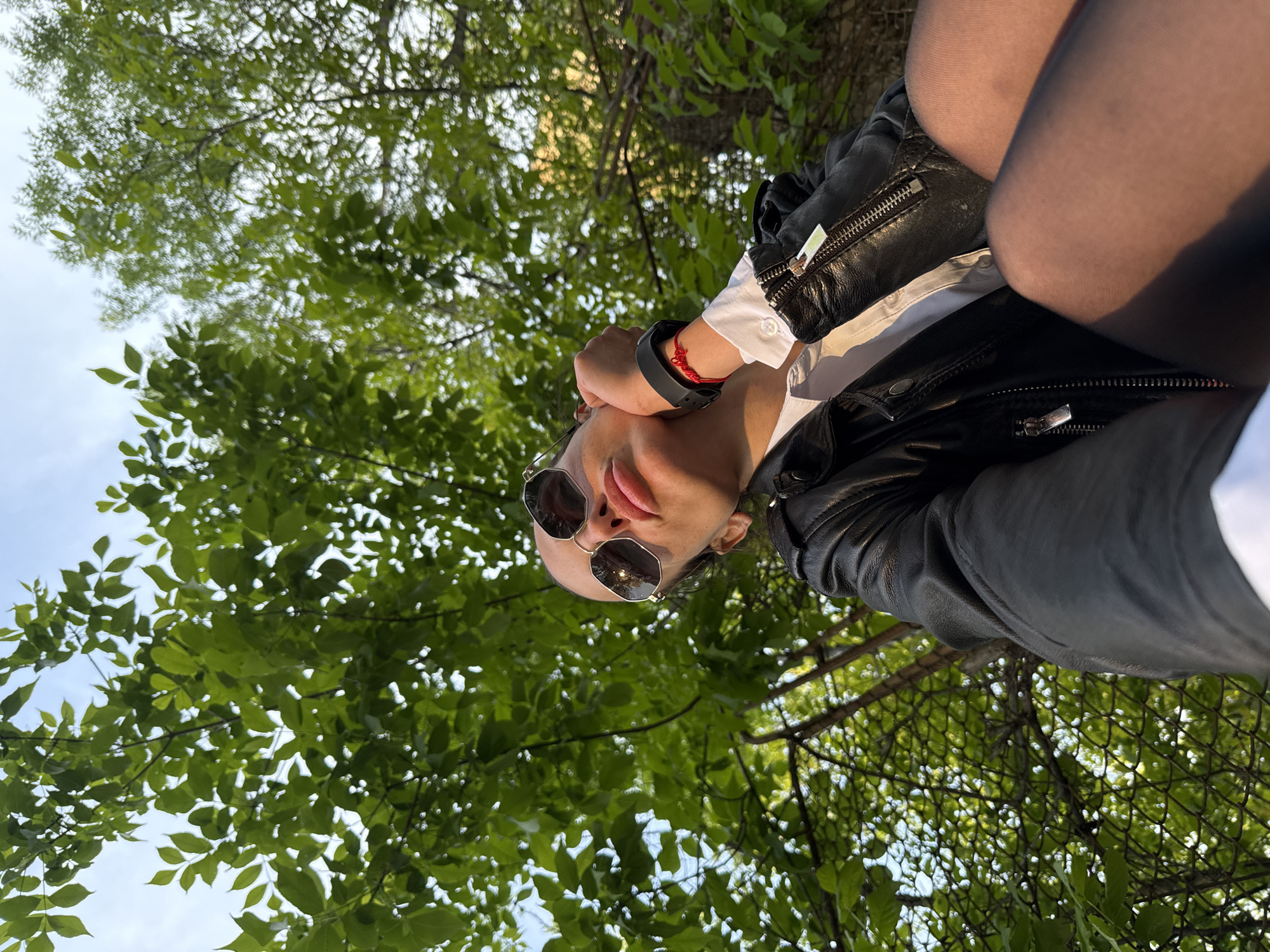 A low-angle shot shows a person wearing sunglasses, a white shirt, a black leather jacket, and black tights. The person is positioned against a backdrop of green foliage and sky. The location appears to be outdoors, possibly a park or wooded area. The city is Pleven, Bulgaria, according to the prompt. The camera is angled upwards toward the person's face. A wire fence is partly visible.