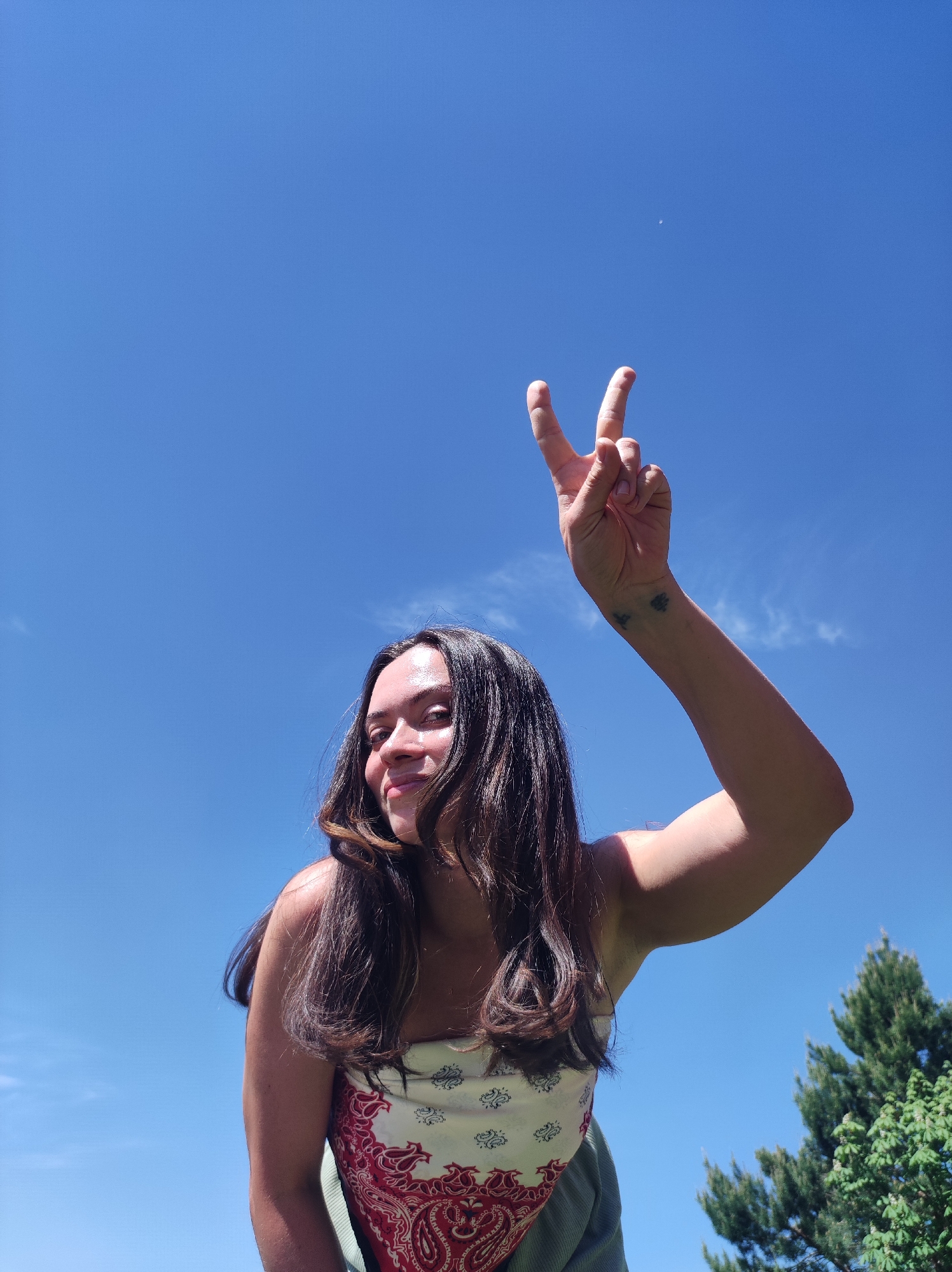 A low-angle shot shows a woman in an outdoor setting under a bright blue sky with a few wispy clouds. She's making a peace sign with her right hand. She has long, dark brown hair and is wearing a white and red paisley patterned top. A glimpse of a green garment is visible on her lower body. A tree is visible in the lower right corner of the frame. The location is unknown and cannot be determined as being in the city of Фоки, Russia.