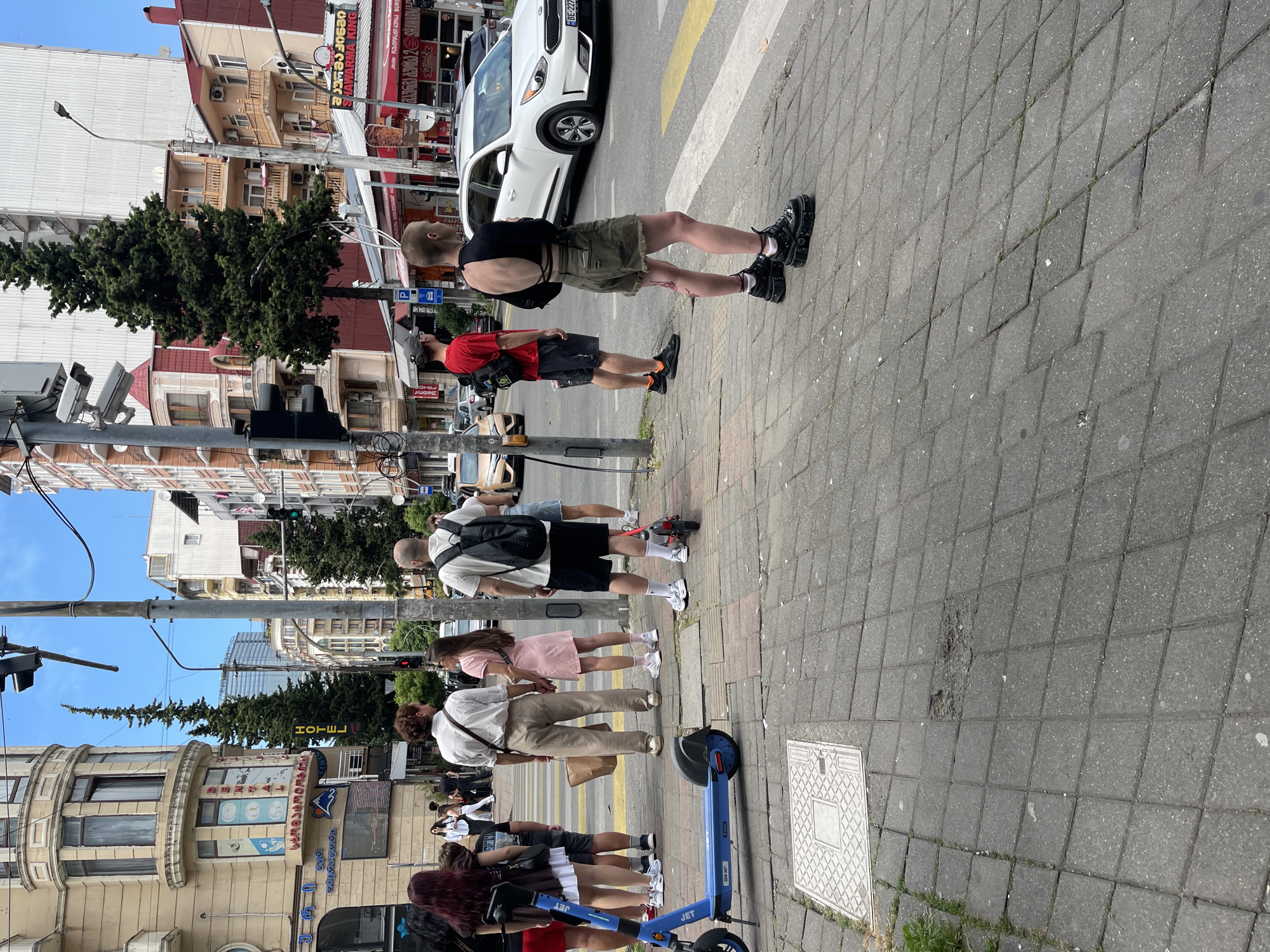 The image captures a street scene in Batumi, Georgia, near a pedestrian crossing. A blue electric scooter is parked on the sidewalk on the left. Several people are visible, some standing and waiting, others walking. In the foreground, a person with a shaved head is wearing a black crop top and olive green shorts, standing in front of a white car. To the left, another person in a red t-shirt and dark shorts is looking forward. Further back, a couple holding hands is visible, with the woman in a pink dress and the man in a light-colored shirt and beige pants. Another person with a backpack is also present. The background shows buildings, trees, traffic lights, and vehicles, indicating a bustling urban environment. The time of day appears to be daytime, with clear sky.