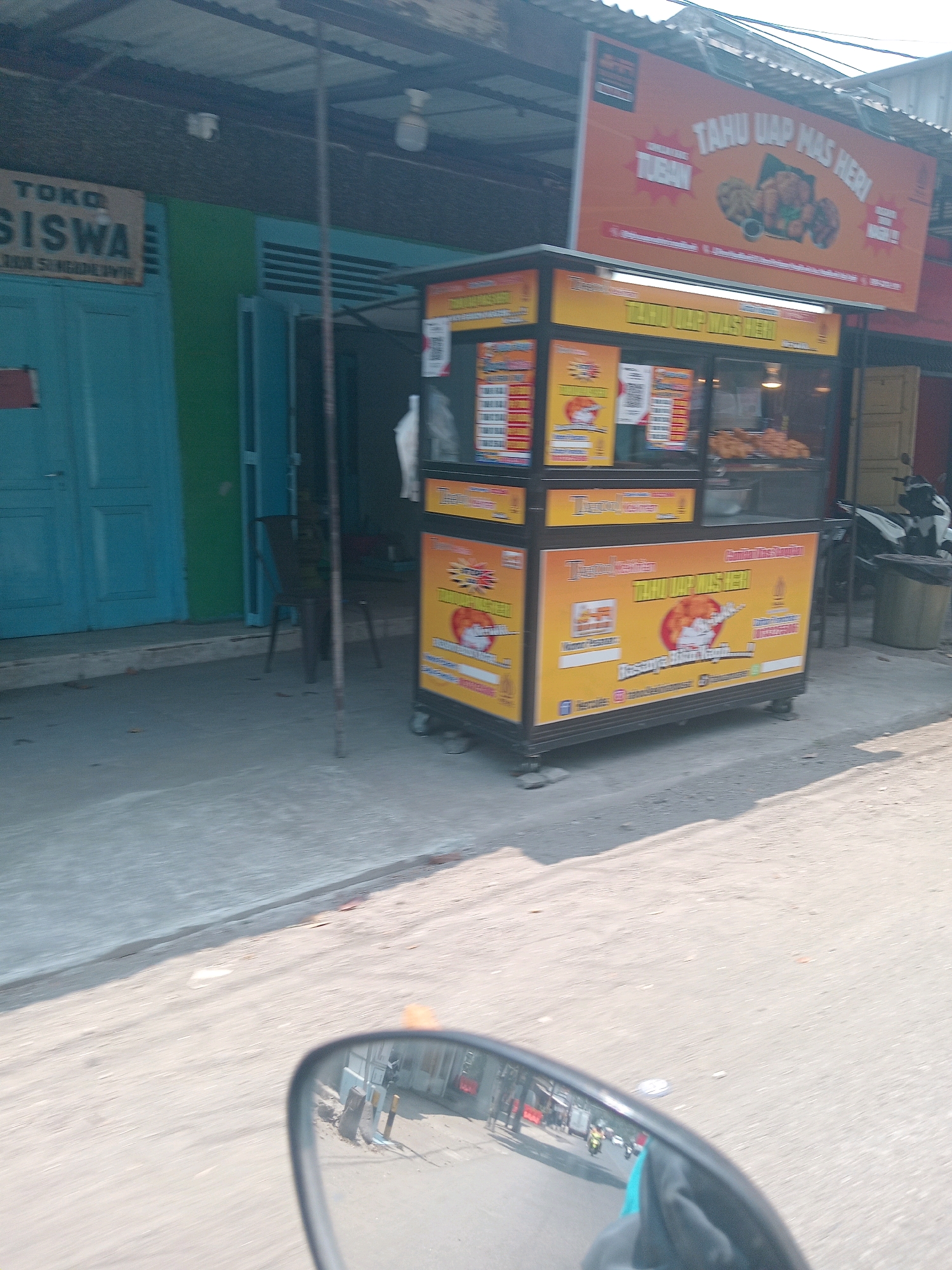 The image shows a street scene in Ngadiluwih, Indonesia. In the foreground, a motorcycle's side mirror reflects a blurry image of the road and surrounding environment. The main subject of the image is a food stall with bright orange and yellow signage that reads "TAHU UAP MAS HERI" and "TUBAH". The stall appears to be selling fried tofu. Behind the stall, there is a building with blue doors and a green facade. Several chairs are visible outside the building, and a trash can is next to a parked motorcycle on the right side of the frame. The ground is paved with concrete, and the sky is overcast. The overall impression is a typical street view of a small business in an Indonesian town.