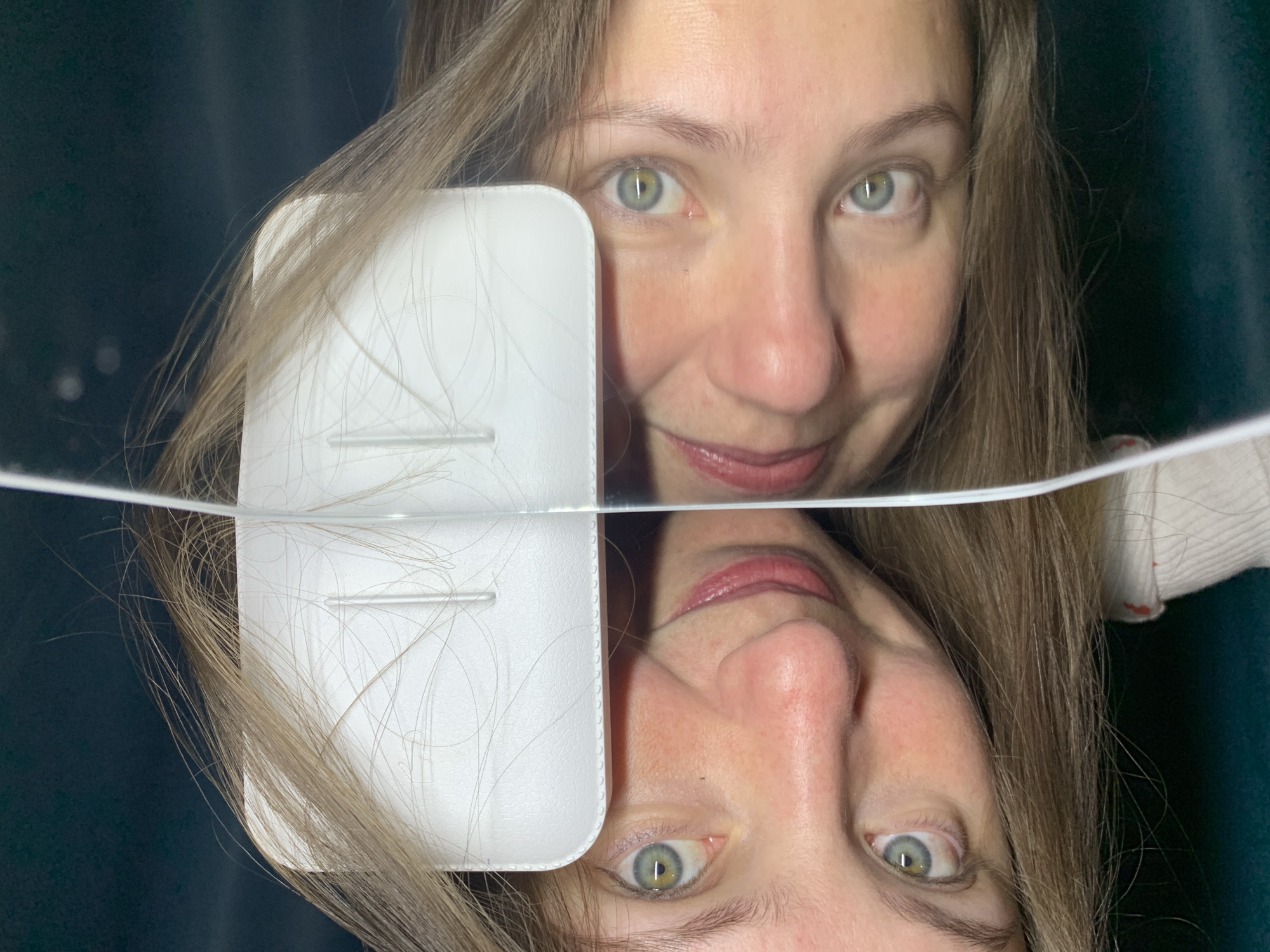 This close-up shot features a woman's face and its reflection, split horizontally by a reflective surface, likely taken in Chaikovsky, Russia. The top half of the image shows the upper part of the woman's face, looking directly at the camera with a gentle smile. She has light-colored eyes (blue-green) and light brown or blonde hair, which cascades around her face and extends onto a white, rectangular object positioned to her left.
The lower half of the image presents an upside-down reflection of the woman's face, clearly showing her nose, mouth, and chin, along with her reflecting hair and eye. Her lips are slightly parted in the reflection, mirroring the smile on her actual face.
A prominent white, rectangular object with two faint horizontal indentations is visible on the left side of the frame. Numerous strands of the woman's long hair are draped across this object, making its precise function unclear, but it appears to be a case or a small device. The background behind the woman is dark, possibly a curtain or wall, providing a simple backdrop. The overall mood is calm, with the woman's expression conveying a pleasant emotion. There is no visible text in the image.