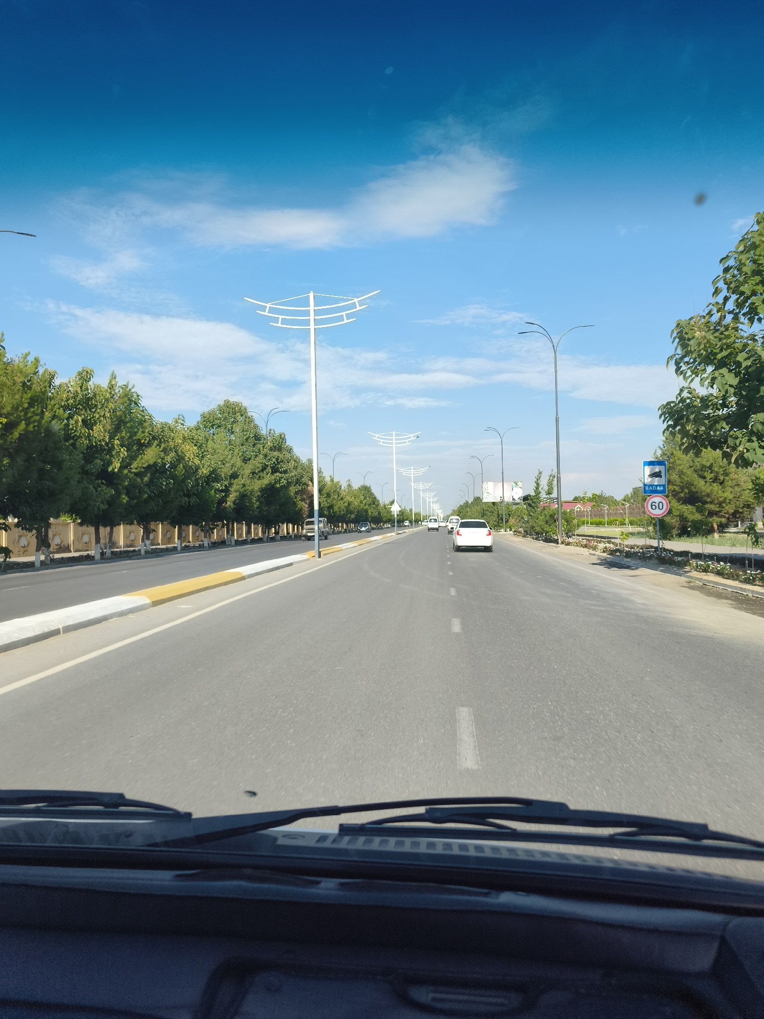 This media file captures a view from inside a vehicle driving down a paved road lined with trees in Buz, Uzbekistan. The road has clear lane markings and a yellow and white striped curb on the left. Tall, modern streetlights with curved tops are visible along the road, suggesting an urban or suburban setting. On the right side of the road, there are more trees and some signage. One sign clearly displays "RADAR" and a circular sign with "60", indicating a speed limit of 60.
Several vehicles are on the road, including a white sedan ahead and another vehicle on the left. The scene appears to be set during the daytime, with a bright blue sky and scattered white clouds, suggesting fair weather. The trees are green and leafy, indicating a warm season. The overall impression is of a routine drive through a well-maintained road infrastructure in a location that appears to be well-developed. There are no people visible within the frame of the road and surrounding environment. The emotion conveyed is one of normalcy and travel.