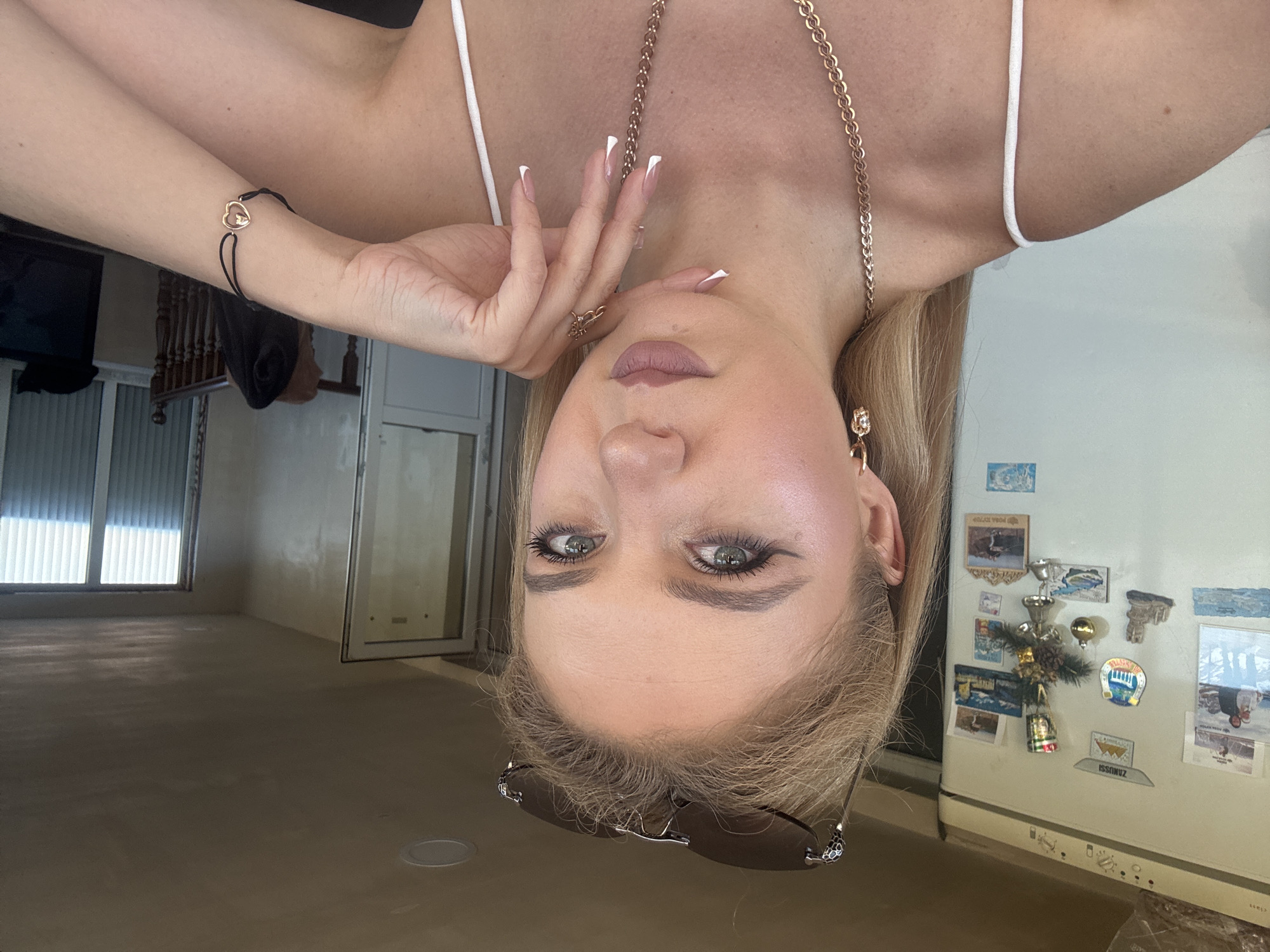 The media file is a photograph of a woman taken from a low angle, causing the image to be upside down. She has blonde hair, green eyes, and is wearing makeup, including lipstick and eyeliner. She is also wearing a white top, a gold necklace, a bracelet with a heart charm, and earrings. Her fingernails are painted with a French manicure. She is holding her left hand up to her face. In the background, there is a refrigerator adorned with magnets, a window with blinds, and a doorway. The scene appears to be indoors, possibly in a kitchen or living area, with neutral-colored walls and flooring. The lighting suggests it could be daytime. There is no discernible text visible in the image.