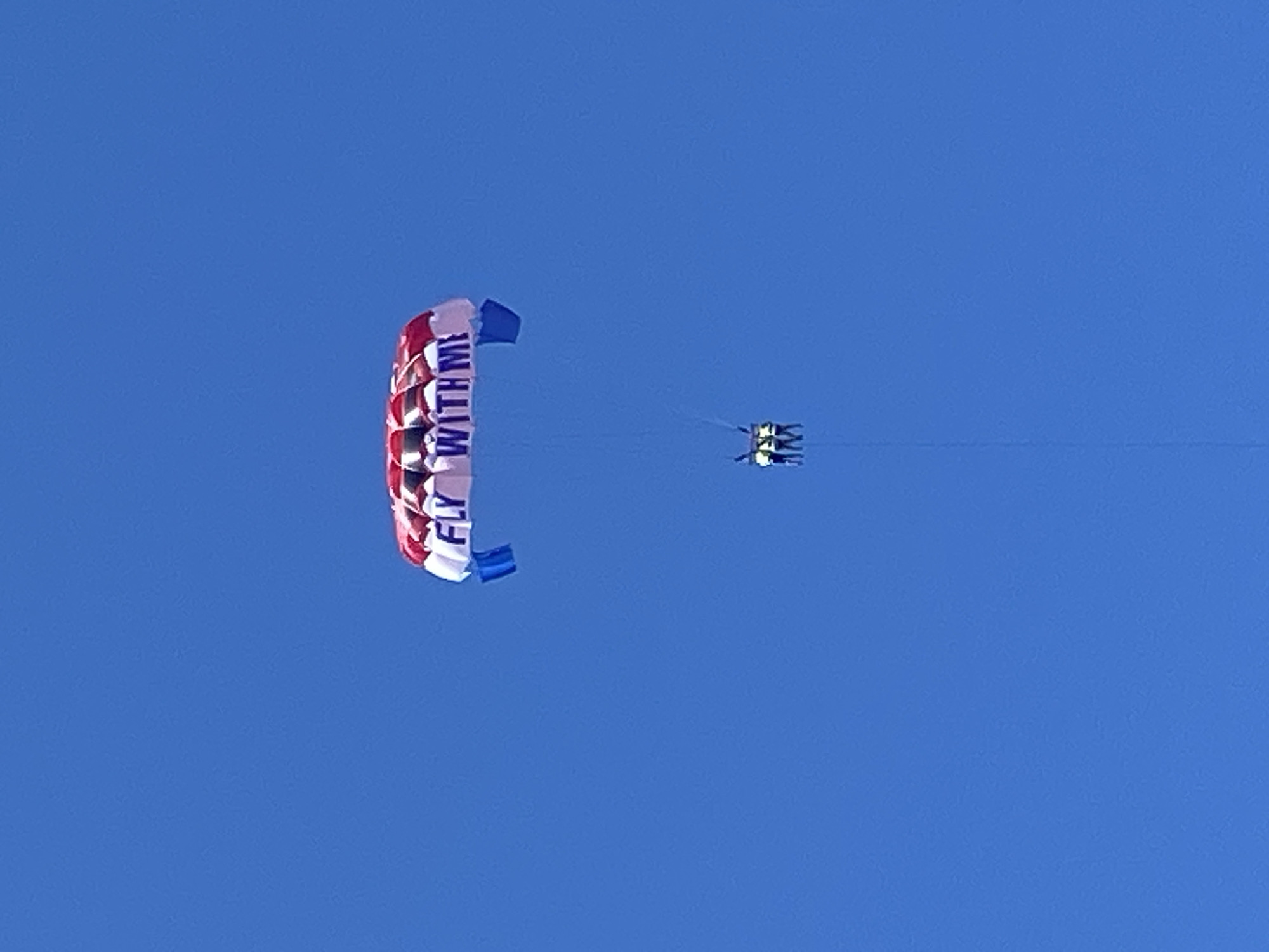 The image captures a parasailing activity against a vast, clear, and uniform deep blue sky. Dominating the upper center of the frame is a large parasail canopy, which features a prominent red and white checkered pattern on its main surface, flanked by solid blue sections on the sides. In the center of the white section, the phrase "FLY WITH ME" is printed in dark, capital letters. Suspended directly beneath the parasail by visible lines are three individuals, harnessed together side-by-side. They appear as small, indistinct figures, wearing light-colored tops and safety gear. A thin tow rope extends downwards from their harness, exiting the bottom of the frame, indicating they are being pulled by a boat not visible in the shot. The scene depicts an aerial view of the parasailing activity, likely occurring over the Adriatic Sea in the vicinity of Budva, Montenegro. No other geographical features, water, or clouds are visible within the frame.