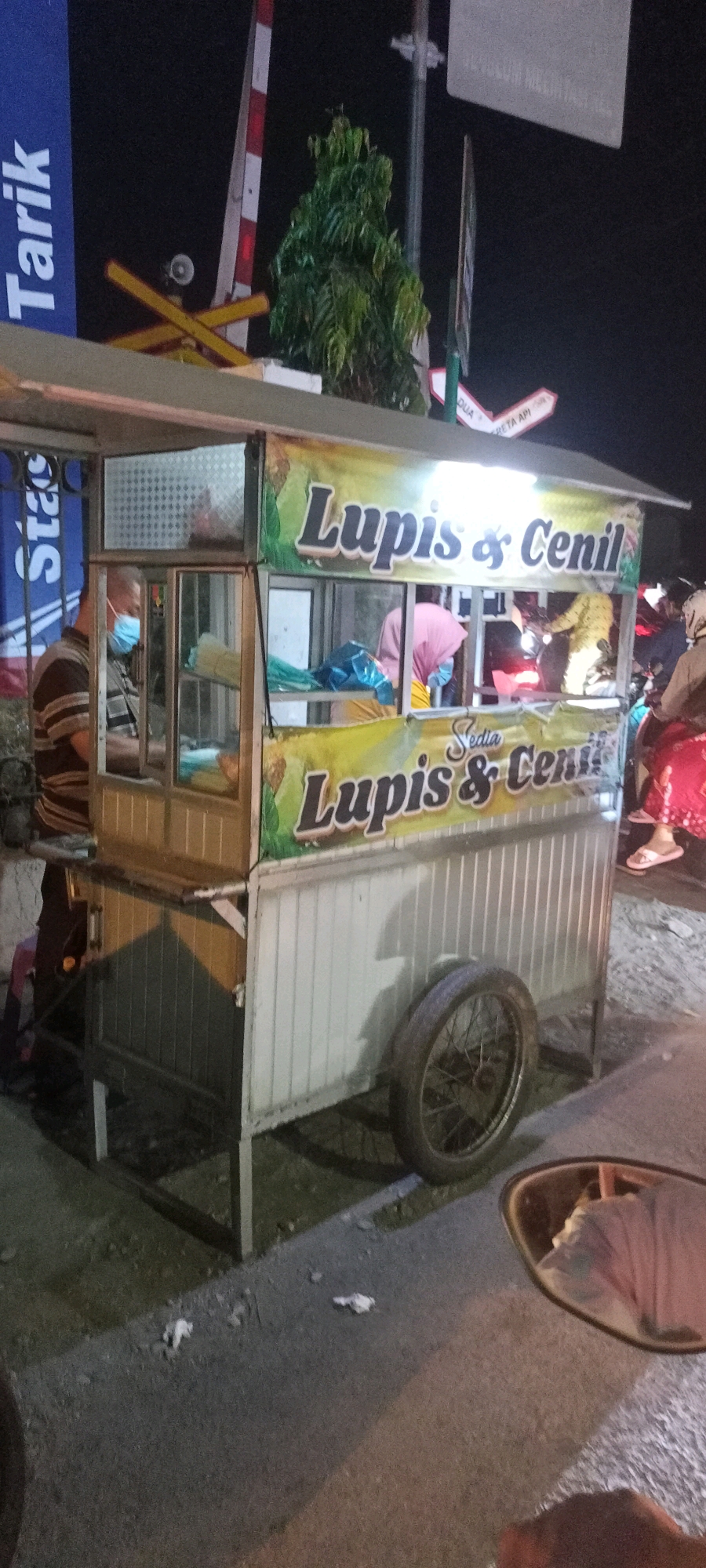An outdoor nighttime scene in Singogalih, Indonesia, features a food cart selling "Lupis & Cenil." The cart, made of metal and wood with clear glass panels, is illuminated and displays colorful banners. A man wearing a striped shirt and a blue face mask is visible inside the cart, possibly a vendor. Another person, wearing a pink headscarf and a pink shirt, is also inside the cart, behind a counter. The cart has a wheel on the right side and is parked on a paved surface, possibly a roadside. In the background, a railway crossing barrier with red and white stripes is visible, along with a tree and some signs. The overall atmosphere suggests a busy street-side food stall at night.