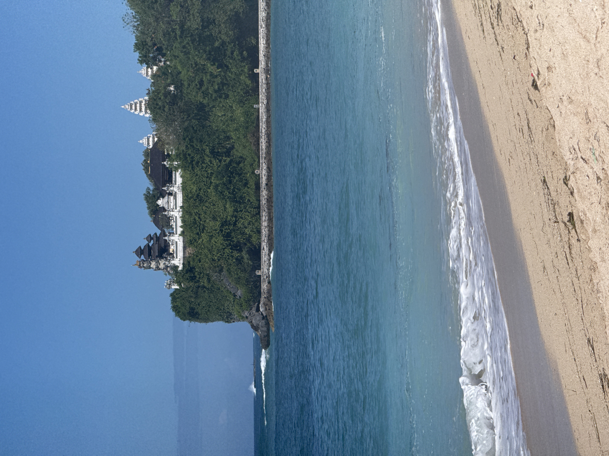 The image shows a coastal scene featuring a temple complex built on a cliffside. The temple has multiple tiered roofs and is partially obscured by lush green vegetation. The cliffside is bordered by a stone wall. Below the cliff, the ocean extends into the distance, with waves breaking along a sandy beach. The sky is a clear blue. The image appears to be taken in Bali, possibly Uluwatu Temple.