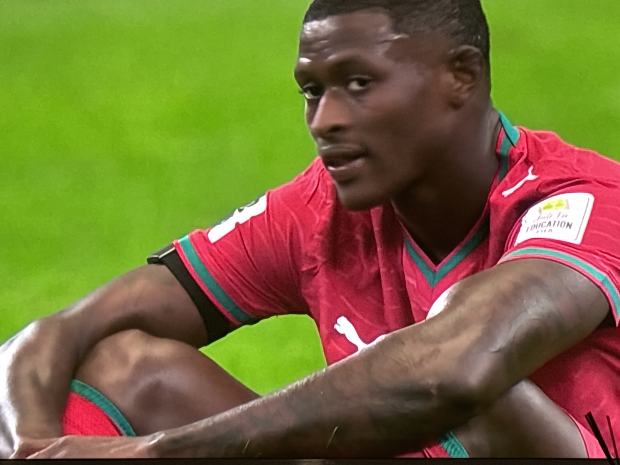 The image is a close-up, angled shot of a Black male football player wearing a red jersey with teal trim and a white Puma logo. He is sitting on a vibrant green surface, possibly a football pitch, and looking off to the side with his mouth slightly open. His arms are bent and crossed over his legs, and his knees are pulled up towards his chest. A patch with text and a logo is visible on his right shoulder. The lighting suggests an outdoor setting during the daytime. There is no indication of the city of Burgas, Bulgaria, in the image.