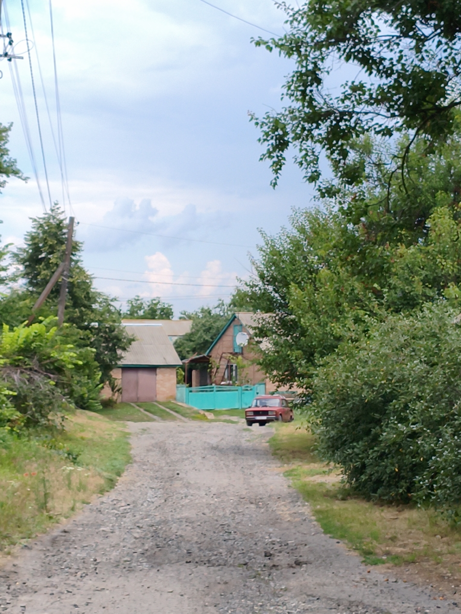 The image depicts a rural scene in the village of Ustyvytsia, Ukraine. A gravel road leads towards several buildings, partially obscured by lush green trees and foliage. On the left side of the road, a wooden utility pole with wires stretches into the distance. Further down the road, a red car is parked. The buildings visible include a brick structure with a large double door and a house with a visible window and a teal-colored fence. The sky is partly cloudy. The environment suggests a warm day.