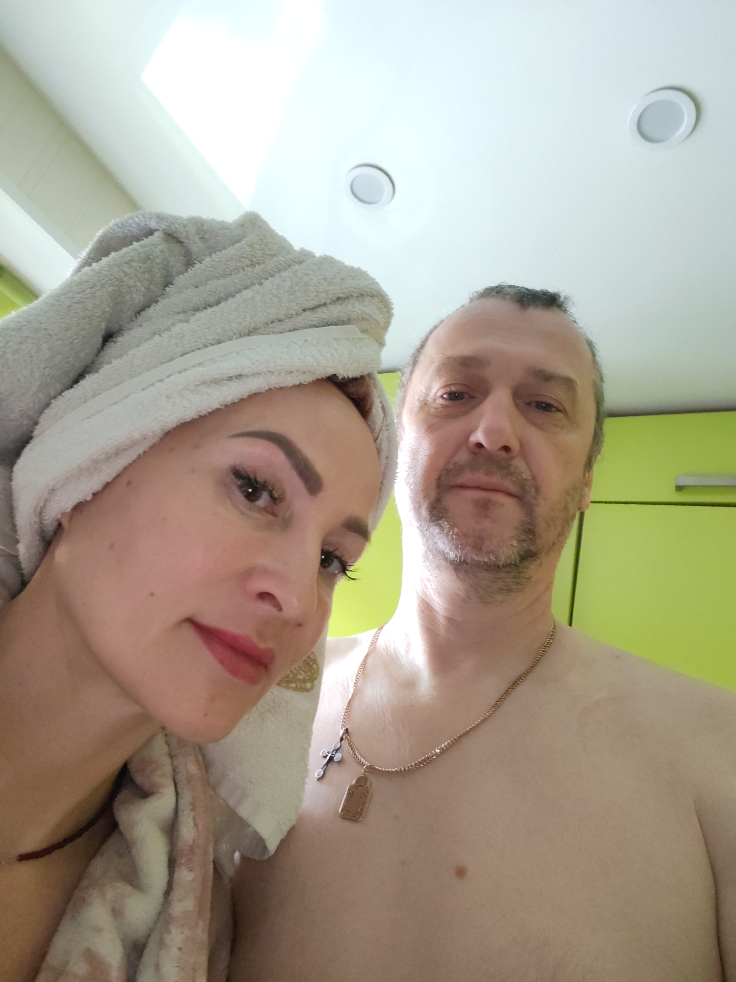 The image depicts a selfie taken indoors, possibly in a bathroom or kitchen, as indicated by the white ceiling lights and light green cabinets. Two individuals are visible: a woman on the left, with a towel wrapped around her head, and a man on the right, who is shirtless and wearing a gold chain with a cross and a rectangular pendant. The woman is wearing red lipstick and has styled eyebrows and eyelashes. The man has a graying beard and appears to be standing close to the woman. There's a possible light source coming from above, near the top left corner. The location is indicated as the city of Chaikovsky, Russia, but there are no specific details in the image to verify this location.