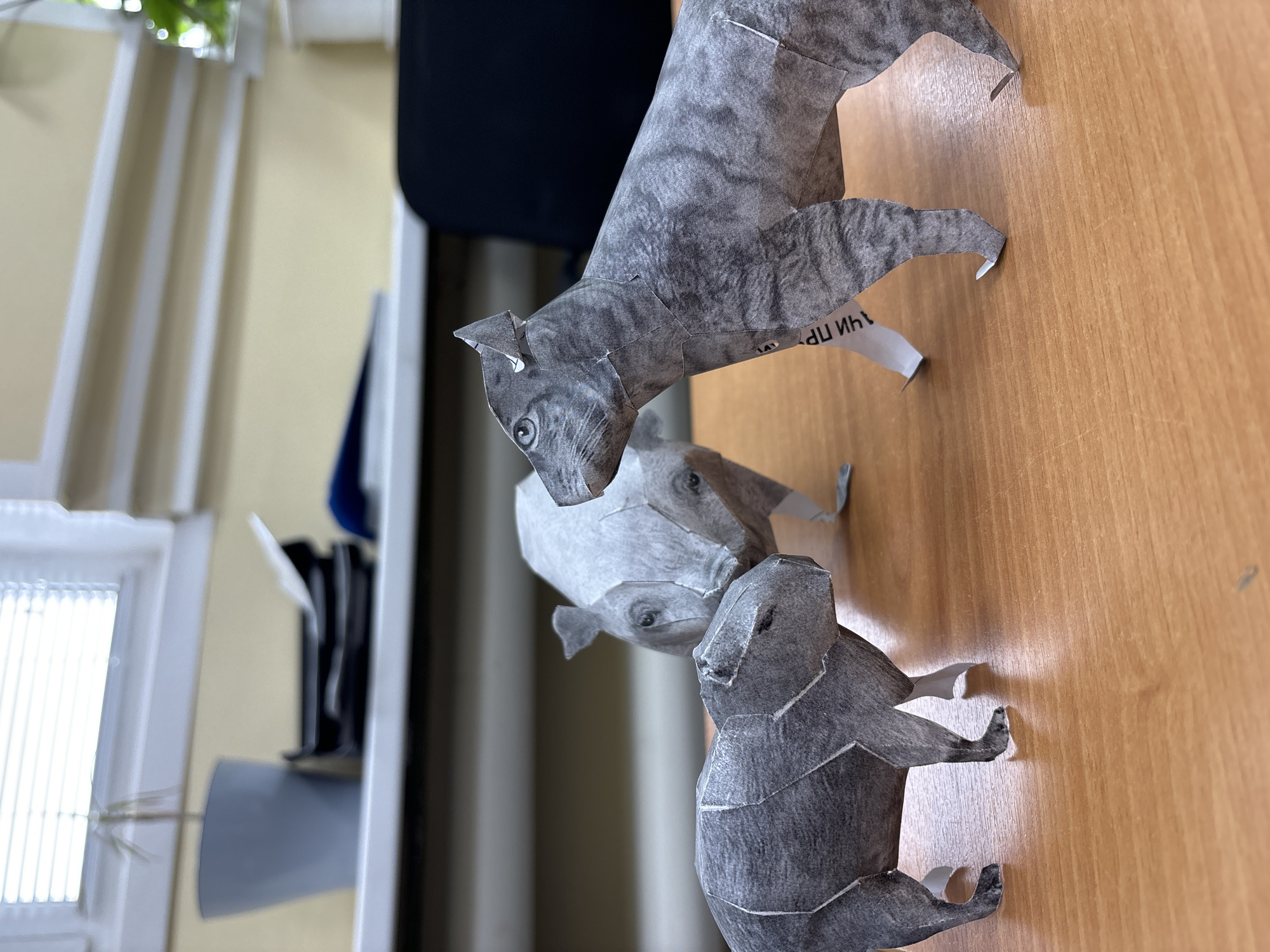 The image shows three paper craft animals on a light brown wooden surface. The top animal is a grey, four-legged creature with visible eyes and a tail. The middle animal, directly below the first, is a lighter grey and also features eyes. The bottom animal is a dark grey and appears to be sitting or squatting. The wooden surface seems to be a desk or tabletop.
The presence of the text "ЧАИ ПР" on a small white piece of paper suggests a potential link to the city of Chaikovsky, Russia, as "ЧАИ" could be an abbreviation for "Chaikovsky."