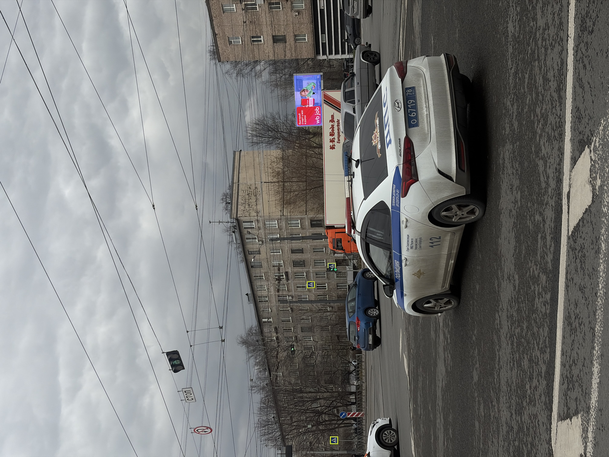 The image shows a street scene in Saint Petersburg, Russia, under a cloudy sky. A white police car is parked on the right side of the frame. The license plate reads "O 6719 78". The car has markings "ПОЛИЦИЯ" (Police) and "112" on its side. Behind it is a blue car. In the background, there are apartment buildings, a billboard, and overhead power lines. A traffic light is also visible.
