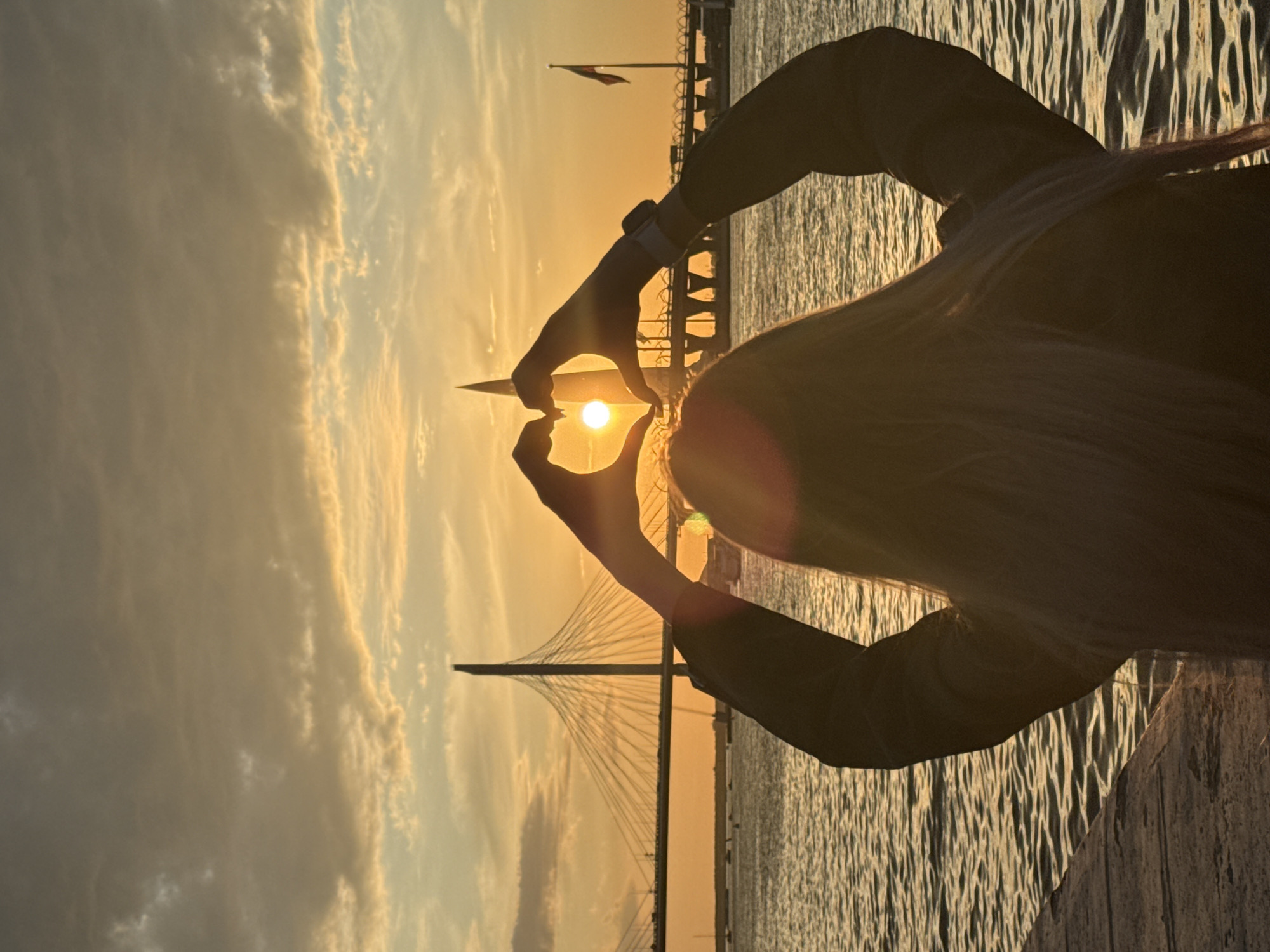This image captures a serene sunset scene in Saint Petersburg, Russia, characterized by warm golden light. The primary subject is a person, likely female with long blonde hair, viewed from behind, standing on what appears to be a pier or dock. Their arms are raised, and their hands are silhouetted against the sun, forming a heart shape that frames the bright, setting sun. A wristwatch is visible on the person's left wrist.
The time of day is clearly sunset, as indicated by the intense golden and orange hues dominating the sky and reflecting off the calm, rippling body of water in the foreground and mid-ground. In the background, a distinctive cable-stayed bridge, consistent with the Western High-Speed Diameter bridge in Saint Petersburg, spans across the water, its structure and cables creating a striking silhouette against the colorful sky. Further in the distance, a flagpole with a flag is visible, along with other faint shoreline structures. The sky features scattered clouds, enhancing the vibrant sunset colors. The overall atmosphere conveys a sense of warmth, beauty, and possibly love or appreciation for the scenic view. No visible text is present in the image.