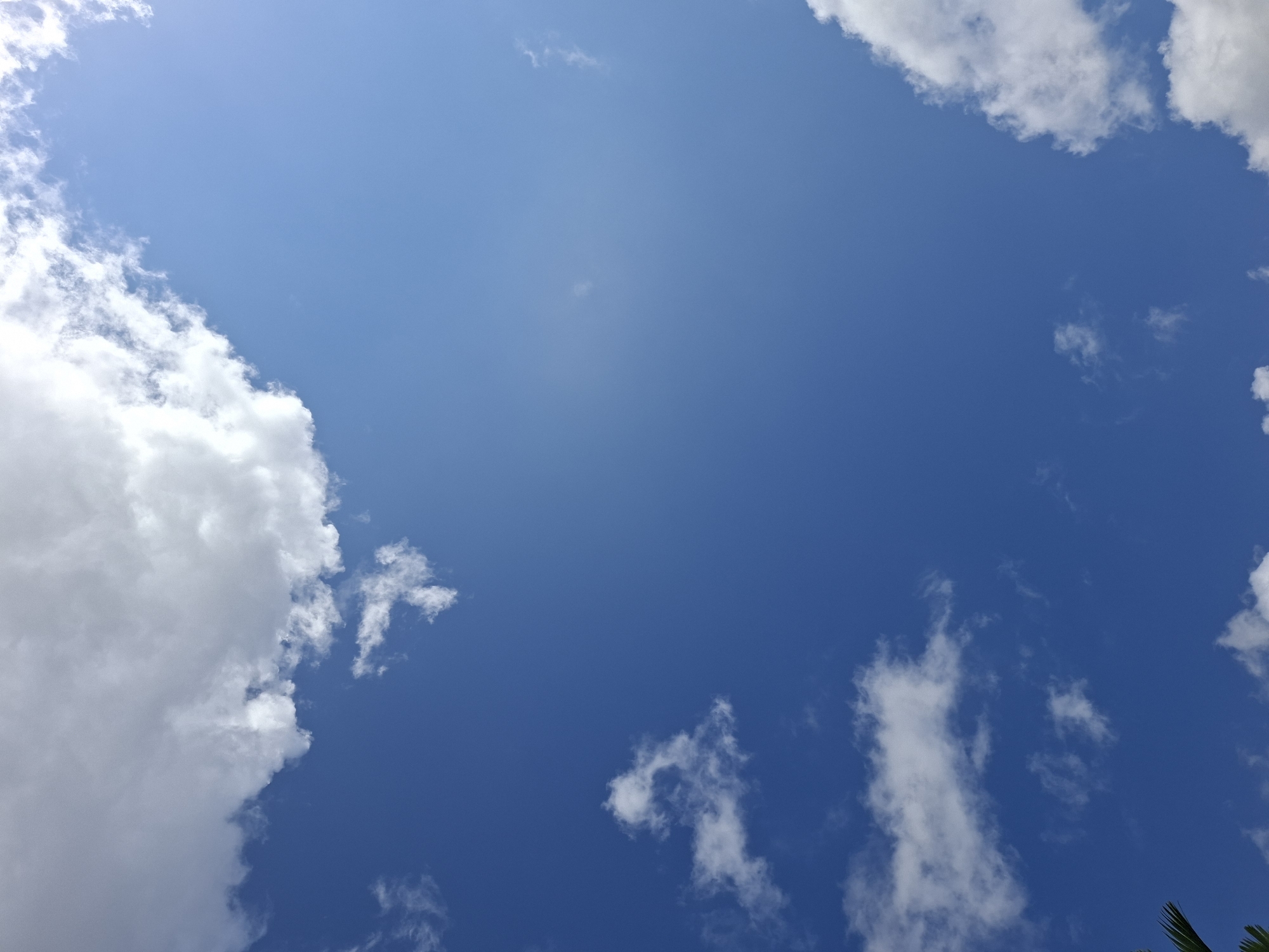 The image captures a view of the sky in Ubud District, Indonesia. The sky is predominantly blue, with varying shades indicating clear areas and areas with cloud cover. White, fluffy clouds are scattered throughout the frame, concentrated mostly along the edges and corners. In the lower right corner, hints of green foliage are visible, suggesting the presence of trees or vegetation. There are no people or man-made structures visible in the frame.