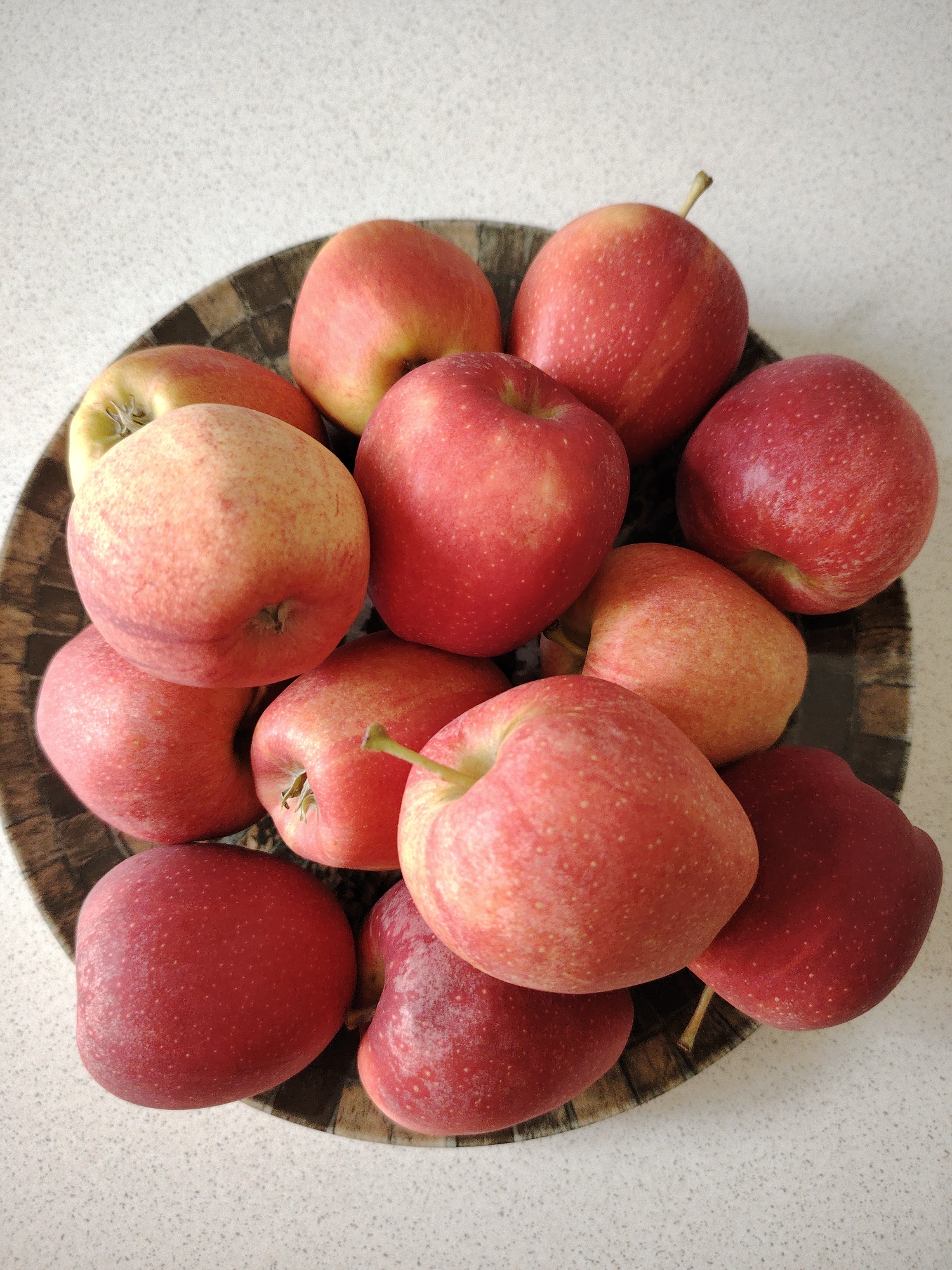 A shallow, dark brown, woven bowl contains a pile of red and pinkish-yellow apples, viewed from directly above. The apples are clustered tightly, with some stacked on top of others. The bowl sits on a speckled, light-colored countertop. The lighting is bright, suggesting daytime, with no indication of weather conditions. The scene is a still life of fruit, likely in a kitchen or dining area. No people, text, or specific events are depicted.