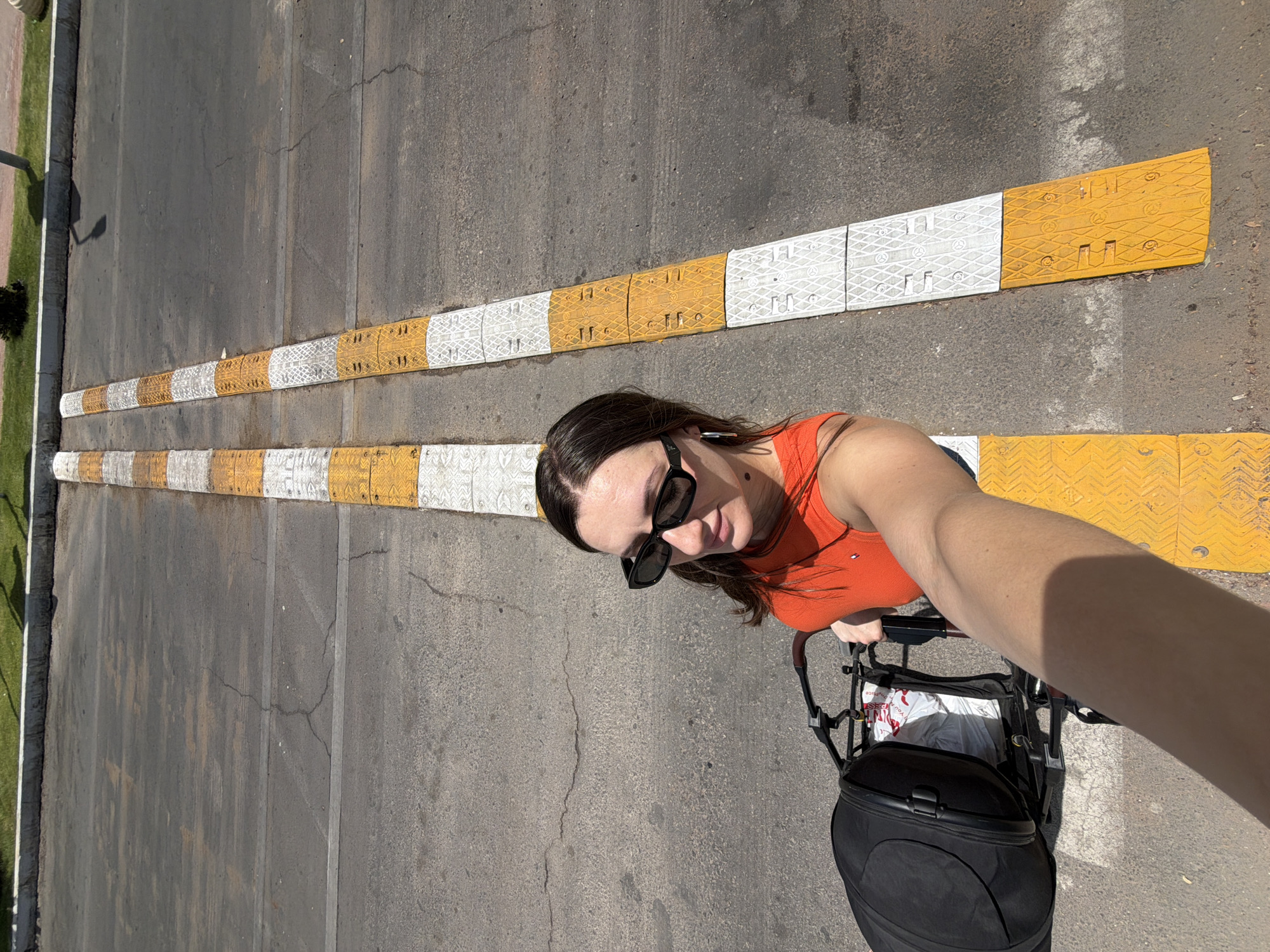 A woman with dark hair and sunglasses is taking a selfie while pushing a stroller with a black bag. She is wearing an orange top and is standing on a cracked asphalt road. Two speed bumps with alternating yellow and white sections are visible on the road. The scene appears to be in Sharm El Sheikh, Egypt, judging by the road markings and surrounding vegetation. The lighting suggests it is daytime and sunny.