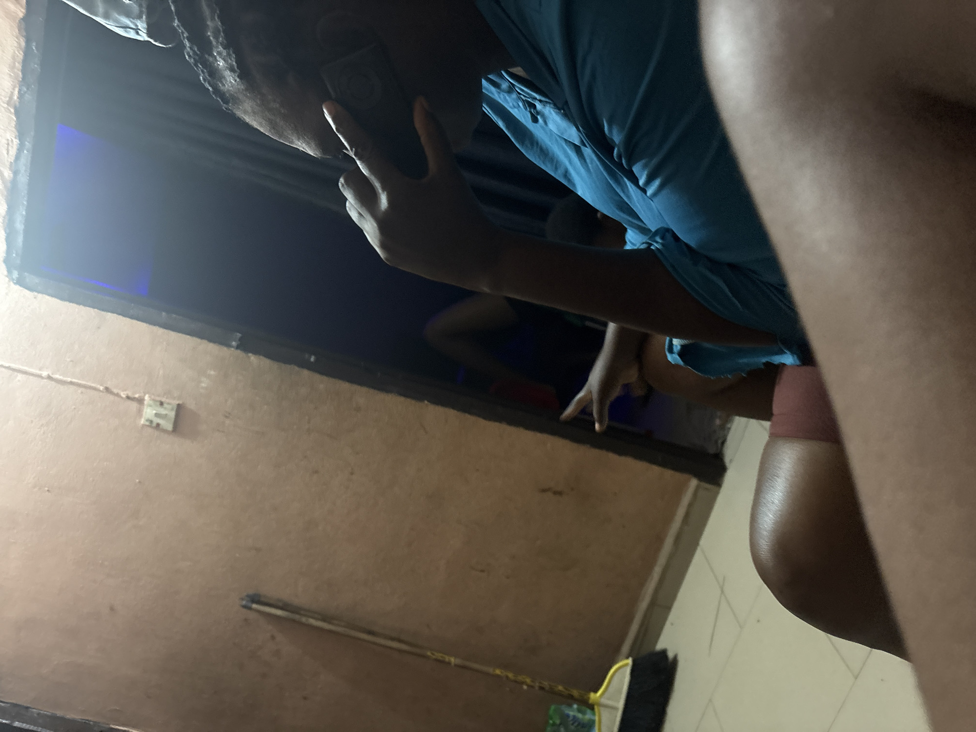 In a dimly lit interior, a person with dark skin, wearing a teal shirt, is seen from the side, holding a dark-colored mobile phone to their ear. Their right hand is raised to their head, with fingers extended around the phone. In the background, another individual's legs are visible, lying down, suggesting a relaxed or resting posture. To the left, a broom with a yellow handle and a white base stands against a textured, light brown wall. A visible electrical outlet is mounted on the same wall, slightly above and to the right of the broom. The environment appears to be a residential or communal space within Onitsha, Nigeria, characterized by the simple wall finish and the presence of everyday household items. The lighting creates strong shadows and highlights, obscuring some details of the surroundings and the individuals.