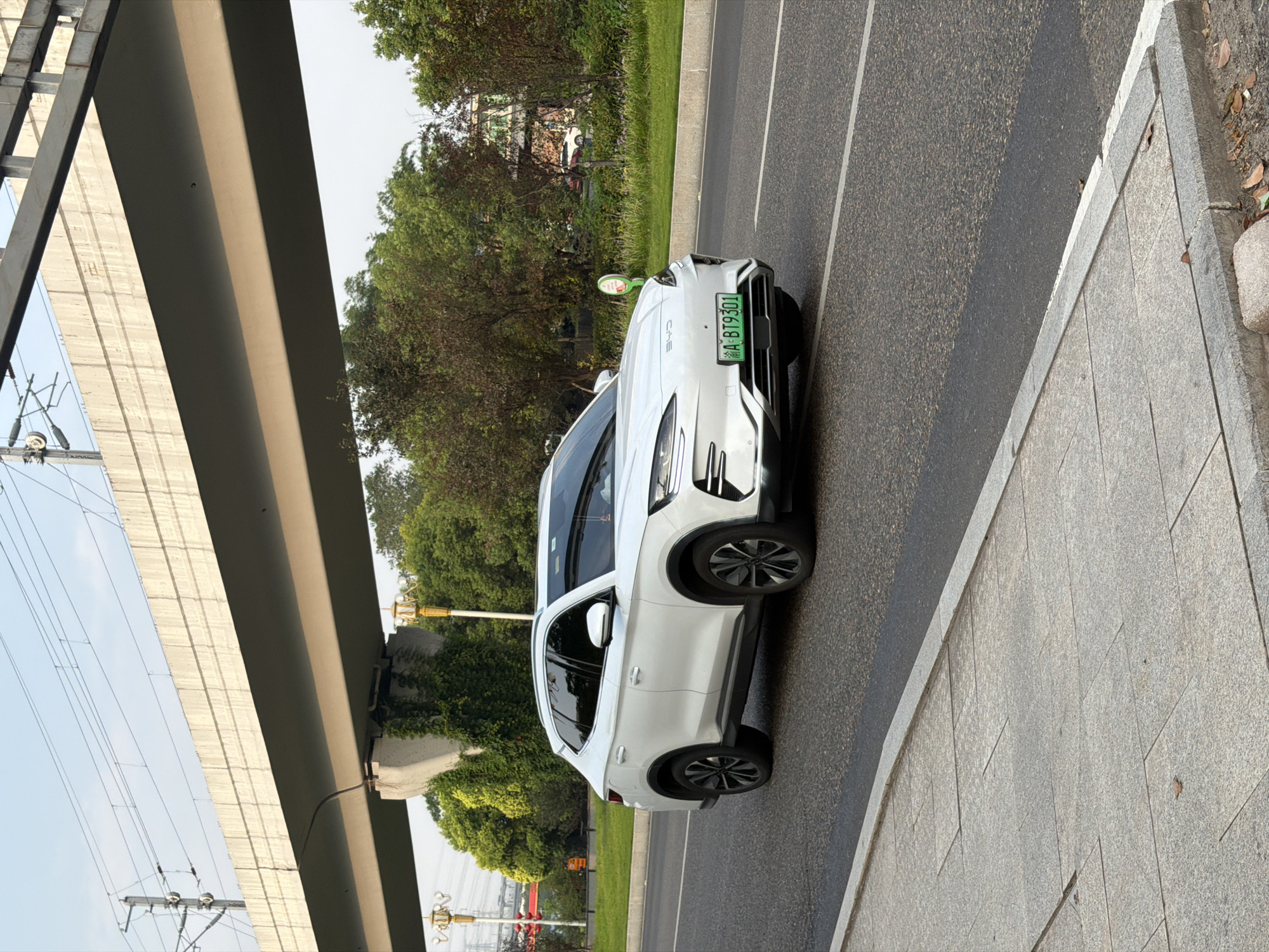 A white BYD electric SUV is seen from a three-quarter front perspective, driving on a paved road in the city of Jiangjin, China. The car's license plate, which is green, reads "A BT9301". The vehicle is positioned in the right lane of the road. To the left of the car, a concrete sidewalk is visible, composed of large grey square tiles. Beyond the road and sidewalk, a grassy median separates the lanes, bordered by trees. In the background, an elevated structure, likely a highway or a railway line, is supported by concrete pillars overgrown with vines. Overhead, power lines are suspended. The sky is clear and a light blue. The lighting suggests it is daytime, possibly late afternoon, as evidenced by the shadows and the warm tones of the light. No people are visible in the frame.
