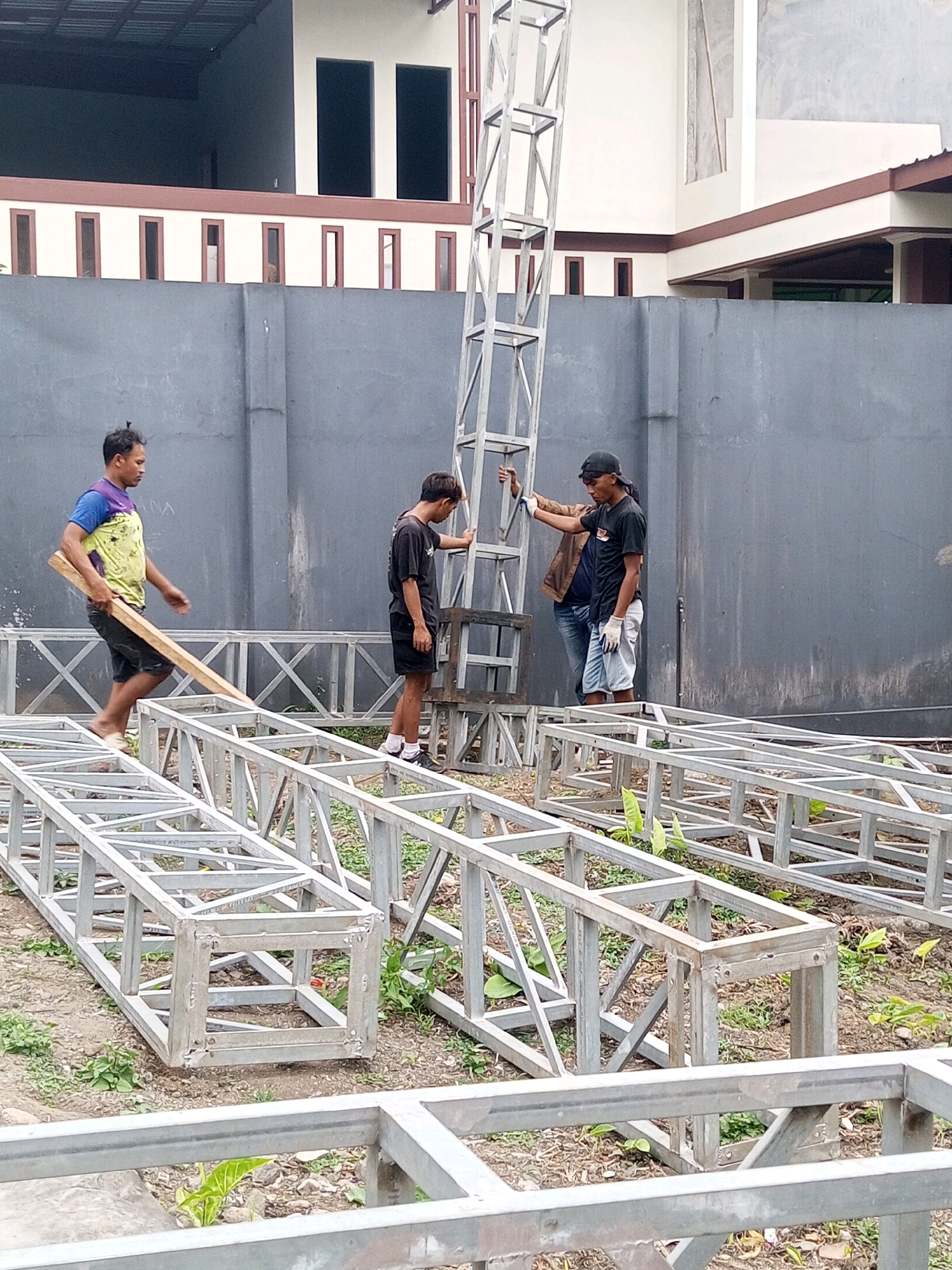 In Andongsili, Indonesia, four individuals are working on constructing a metal truss structure. Two men are actively engaged with the structure, with one carrying a wooden plank and the other assisting with the vertical tower element. A third man is visible in the background, and a fourth, partially obscured, appears to be working alongside. The scene is set outdoors, with a grey wall forming a backdrop and an unfinished building visible behind it. The ground is covered with dirt and sparse vegetation, and several sections of the metal truss lie on the ground, suggesting an ongoing assembly process.