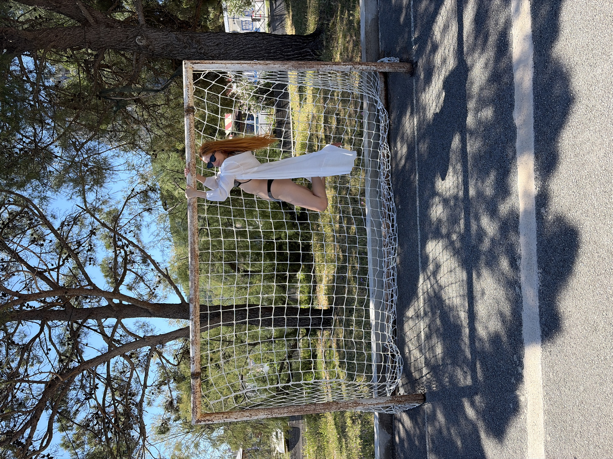 An outdoor, sunlit image captures a woman posing against a metal sports goal, likely in Budva, Montenegro. The woman has long, flowing reddish-brown hair and is wearing dark sunglasses, a black two-piece bikini, and a loose, white long-sleeved cover-up worn open. She is positioned with one leg bent, leaning back slightly and holding onto the white net of the goal frame.
The goal itself appears to be an older model, with visible rust on its metal posts. It stands on a paved surface, likely asphalt or tarmac, which features a distinct white line. Behind the goal, a lush natural environment unfolds, characterized by numerous large trees with dense foliage, appearing to be pine trees, set against a clear blue sky. Bright sunlight casts strong, intricate shadows of the trees and the goal net across the paved ground, indicating a clear, sunny day, most likely during the mid-morning or afternoon. The scene evokes a recreational area, possibly a park or sports court near a coastal region. There is no visible text in the image.