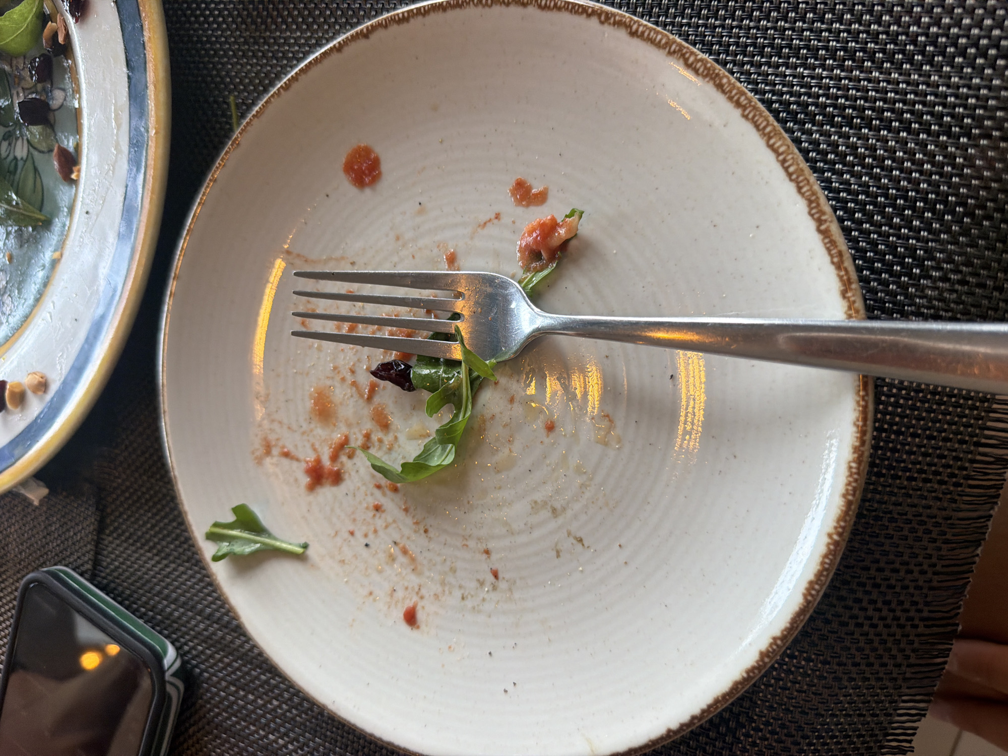 A metal fork rests on a white plate with a brown rim, which is on a dark textured placemat. The plate is dirty with remnants of food, including tomato sauce and a few green leaves. In the background, another plate with food is visible. A smartphone is also on the left side of the image. The setting appears to be a restaurant in Baan Bo Phut, Thailand.