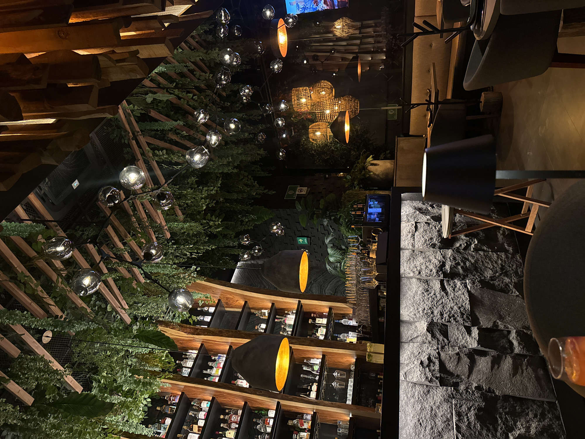 The image depicts the interior of a bar or restaurant in San Bernardino Tlaxcalancingo, Mexico. The setting is dimly lit, with a focus on decorative lighting. Multiple strands of spherical lights hang from the ceiling amidst artificial greenery and wooden beams. Additional lighting is provided by pendant lamps with gold-colored interiors. A well-stocked bar is visible with shelves filled with liquor bottles. The foreground features a textured stone wall and glimpses of furniture, including a table and chairs. There are no people visible in the scene. The overall atmosphere is modern and stylish.