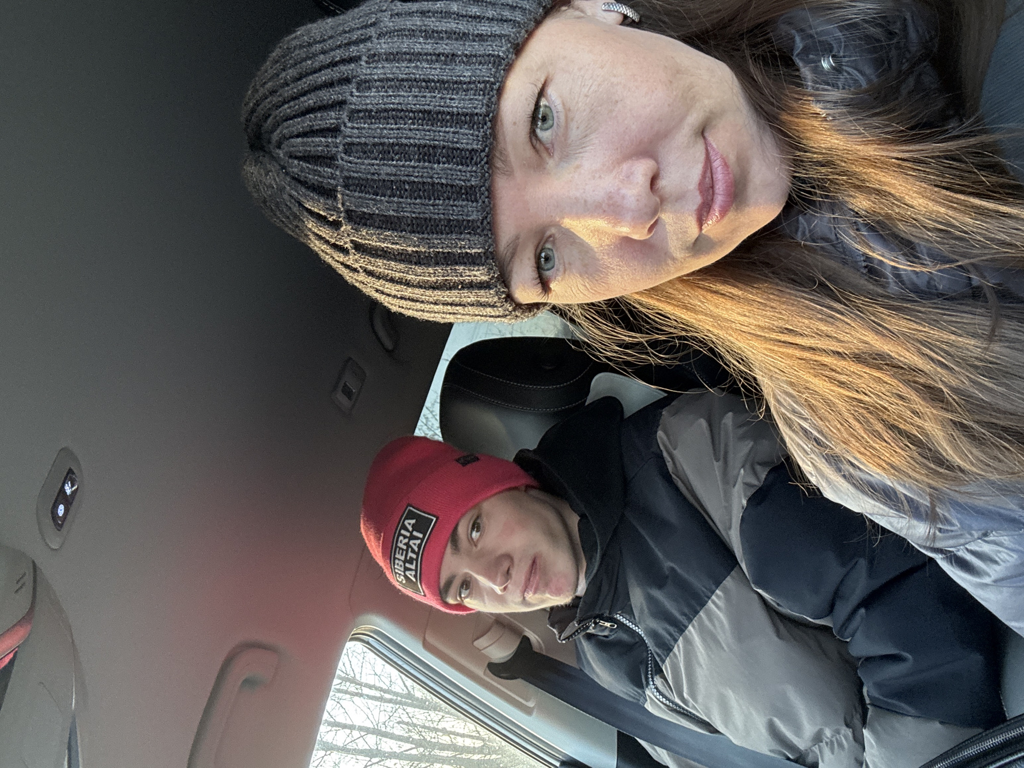 A selfie of two people inside a vehicle. The first person, a woman, is wearing a gray beanie and dark jacket with light brown hair visible. The second person, a younger individual, is wearing a red beanie with a logo "Siberia Altai" and a black and gray jacket, secured with a seatbelt. A blurred view of trees is visible through the window. It is not possible to determine whether this image was taken in the city of Chaikovsky, Russia from the contents of the image.