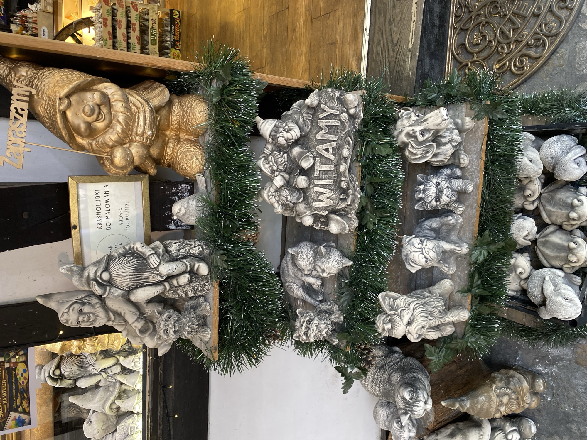 The image displays a multi-tiered wooden retail display stand, adorned with green artificial tinsel garland, located at what appears to be the entrance of a shop on Jatki Street in Wrocław, Poland. This location is identified by the Polish text "NA JATKACH" visible on a sign reflected in the upper left shop window, and the prevalence of gnome statues which are characteristic of Wrocław.
The stand is filled with various unpainted plaster or concrete garden statues:
* **Top Shelf:** Features a grouping of gray unpainted gnome statues, one with a small gray dog statue at its base. To the right, a large golden-painted gnome statue holds a wooden sign with "Zapraszamy" (Welcome) inscribed. A smaller gray bird statue is positioned nearby. A framed sign in the background states "KRASNOLUDKI DO MALOWANIA" and "GNOMES FOR PAINTING".
* **Second Shelf:** Contains a small gray kitten statue and two larger gray cat statues, one appearing to sleep. To the right, a rectangular gray statue features three gnomes above the raised word "WITAMY" (Welcome).
* **Third Shelf:** Displays three gray dog statues: a Yorkshire Terrier, a Pug, and a Basset Hound-like figure.
* **Bottom Shelf/Container:** Holds a collection of smaller gray animal statues, including multiple frogs and rabbits.
The background on the right reveals a shop interior with a light wooden floor and shelves stocked with products, some labeled "UVA Fruit" and "LA RURICA". A decorative bronze-colored metal doormat with an ornate pattern is visible on the ground in the lower right foreground. No people are present in the scene. The items are displayed for sale.